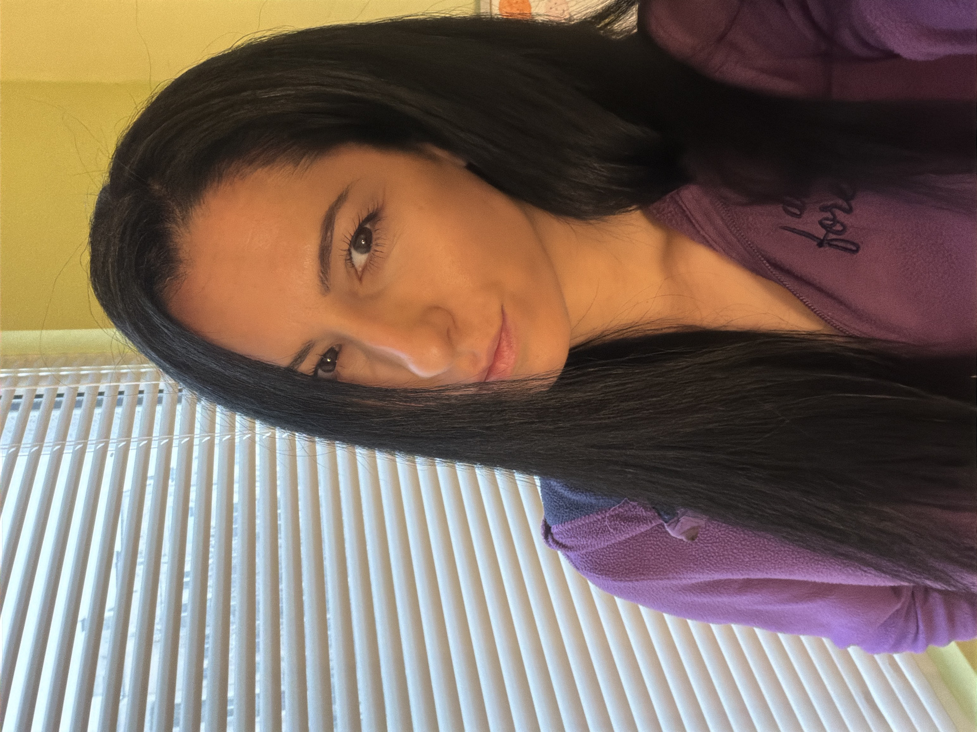 A woman with long, dark hair and light skin is wearing a purple fleece jacket. She is positioned in front of a window with closed white blinds. The background suggests an indoor setting with light yellow walls. It is not possible to determine the location as Pleven, Bulgaria, from the image alone.