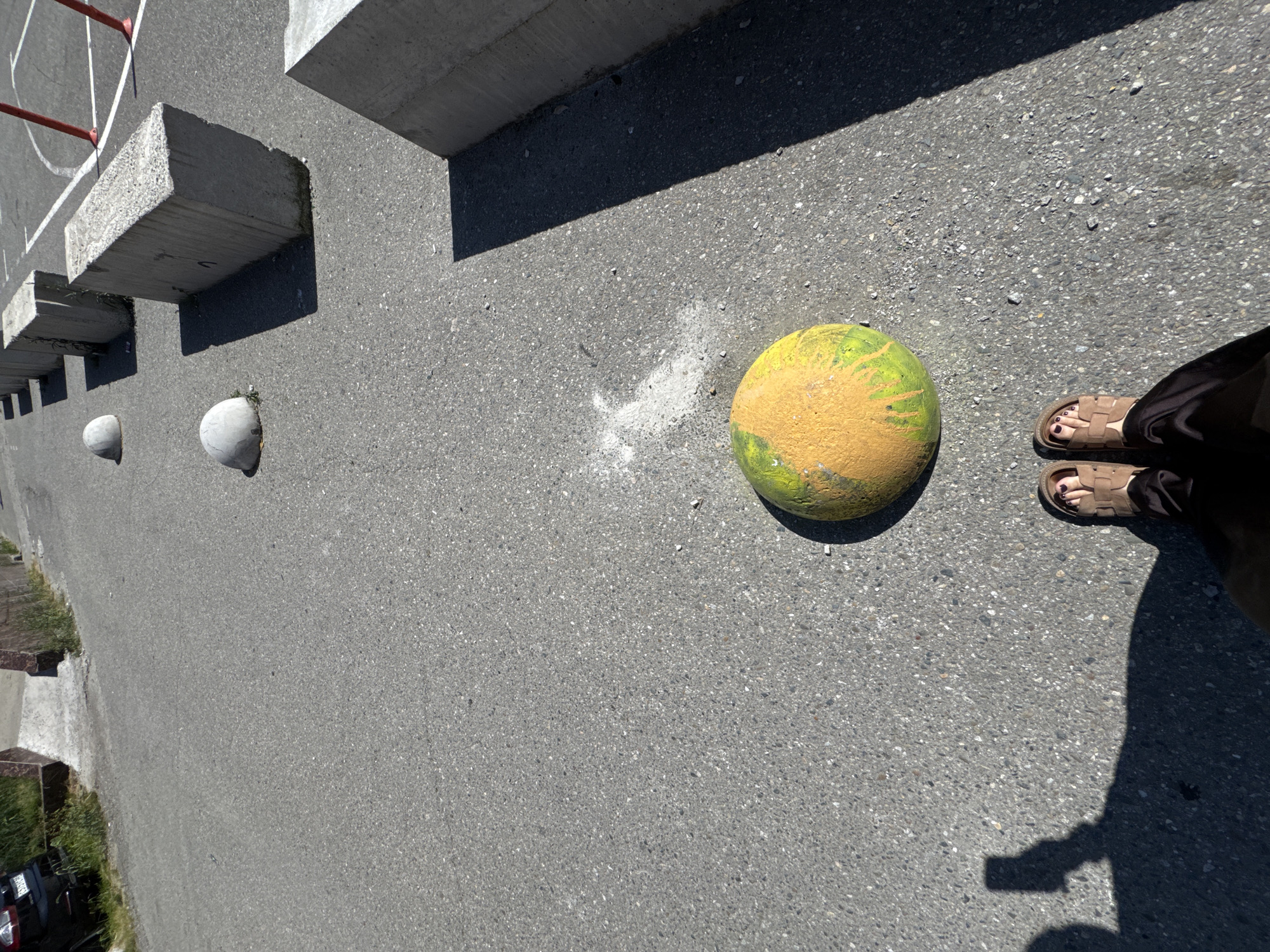 The image provides a ground-level perspective of an outdoor paved area, likely asphalt, illuminated by bright sunlight. A large, roughly spherical object, painted vibrantly in yellow and green resembling a watermelon, occupies the mid-right of the frame, resting on the asphalt. To its immediate left, a small, irregular patch of white dust or debris is visible on the dark gray surface.
In the lower right corner, the feet of a person are visible, wearing brown open-toed sandals. Dark red nail polish is discernible on the person's toes. A long, distinct shadow belonging to the person and possibly an overhead structure extends from the lower right towards the center of the image.
Towards the upper left of the frame, a series of large, rectangular concrete blocks are arranged, casting sharp, elongated shadows diagonally across the asphalt, indicating a sunny day. Below these blocks, two smaller, light gray, domed or hemispherical concrete objects are partially embedded in the ground. In the far upper-left, a red pole, likely from a basketball hoop, and a segment of a white line marking on a sports court are visible. The setting appears to be an outdoor public or recreational space in Sochi, Russia, under clear skies during the day. There is no visible text in the image.