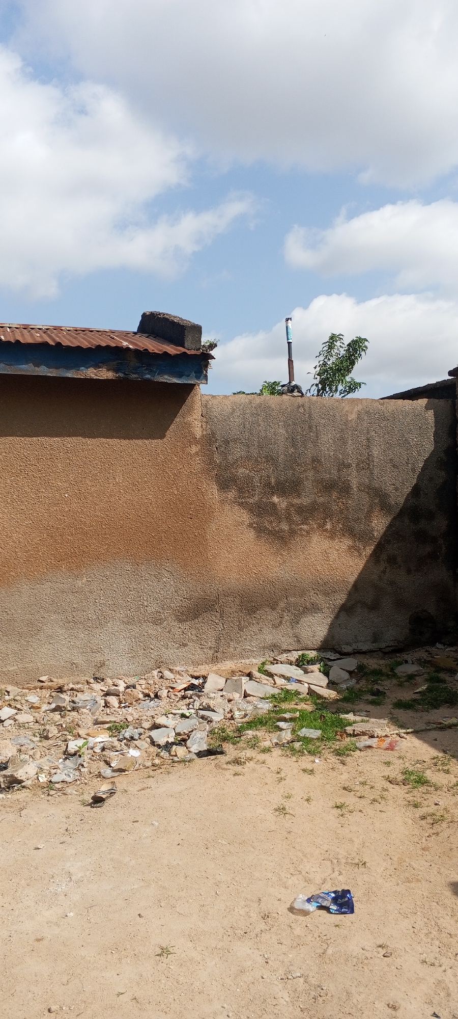 This image depicts an outdoor scene in Jalingo, Nigeria. In the background, a partially visible building with a rusty corrugated metal roof extends from the left side of the frame, supported by a textured, light brown wall. To the right of the building, a tall, slender metal pole with some blue and white banding stands vertically, topped with what appears to be a white container. A small tree with green foliage is situated behind the pole. The wall in the foreground is rough and light brown, with darker, rougher patches and a distinct shadow cast across its right half. The ground in front of the wall is covered with loose rocks, debris, and sparse green grass. A single flip-flop is visible lying on the sandy ground in the middle-left of the image. Further to the right on the sandy ground, a crumpled blue and white plastic bag lies on the ground. The sky above is blue with scattered white clouds.