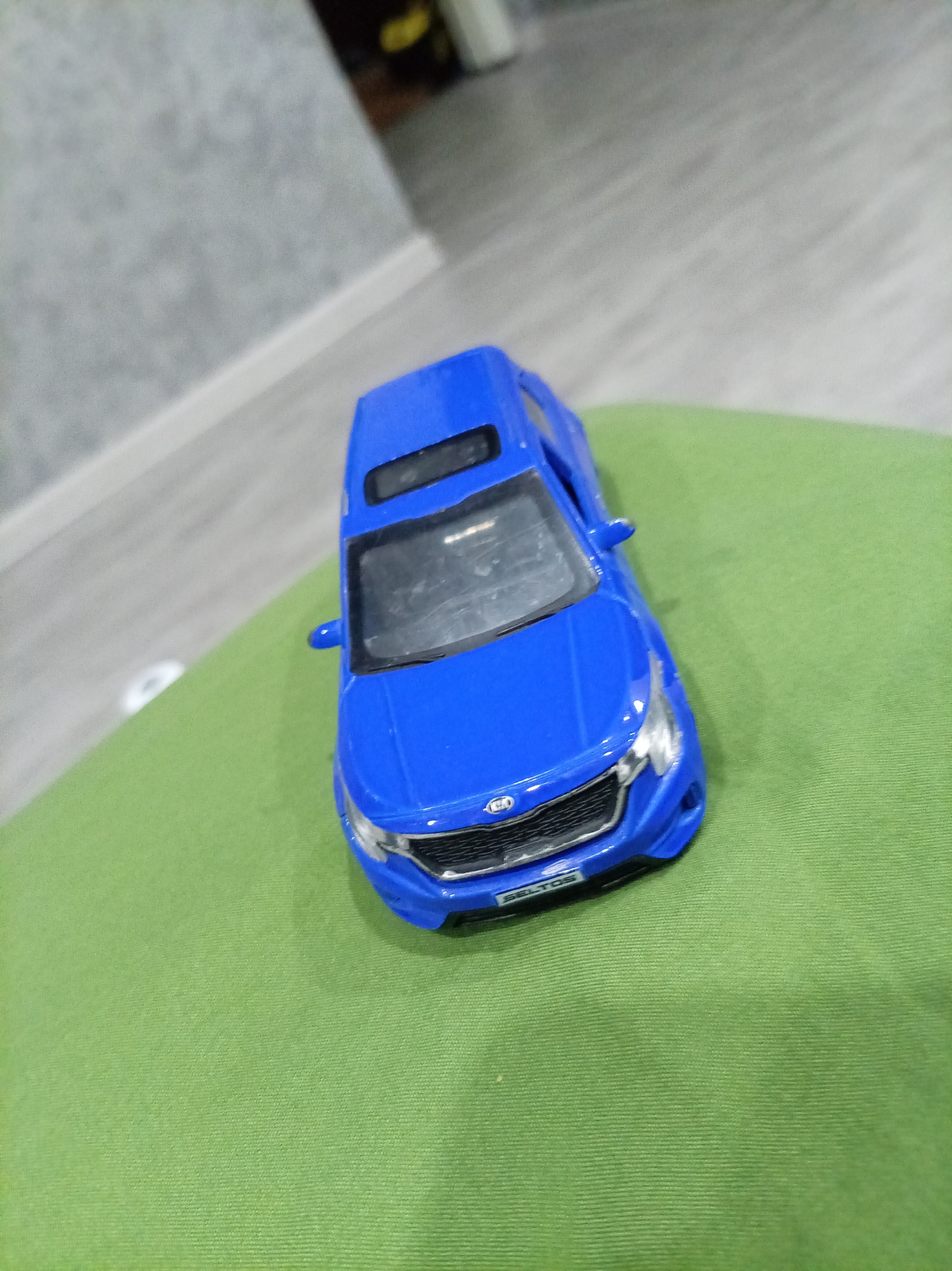 A blue toy car is positioned on a green surface. The car is facing forward and slightly angled to the left. The background is out of focus, showing a gray wall with a white baseboard and a hint of a wooden floor. The scene is likely indoors. There is no indication of the city of Oktyabrsk, Russia, in the image.