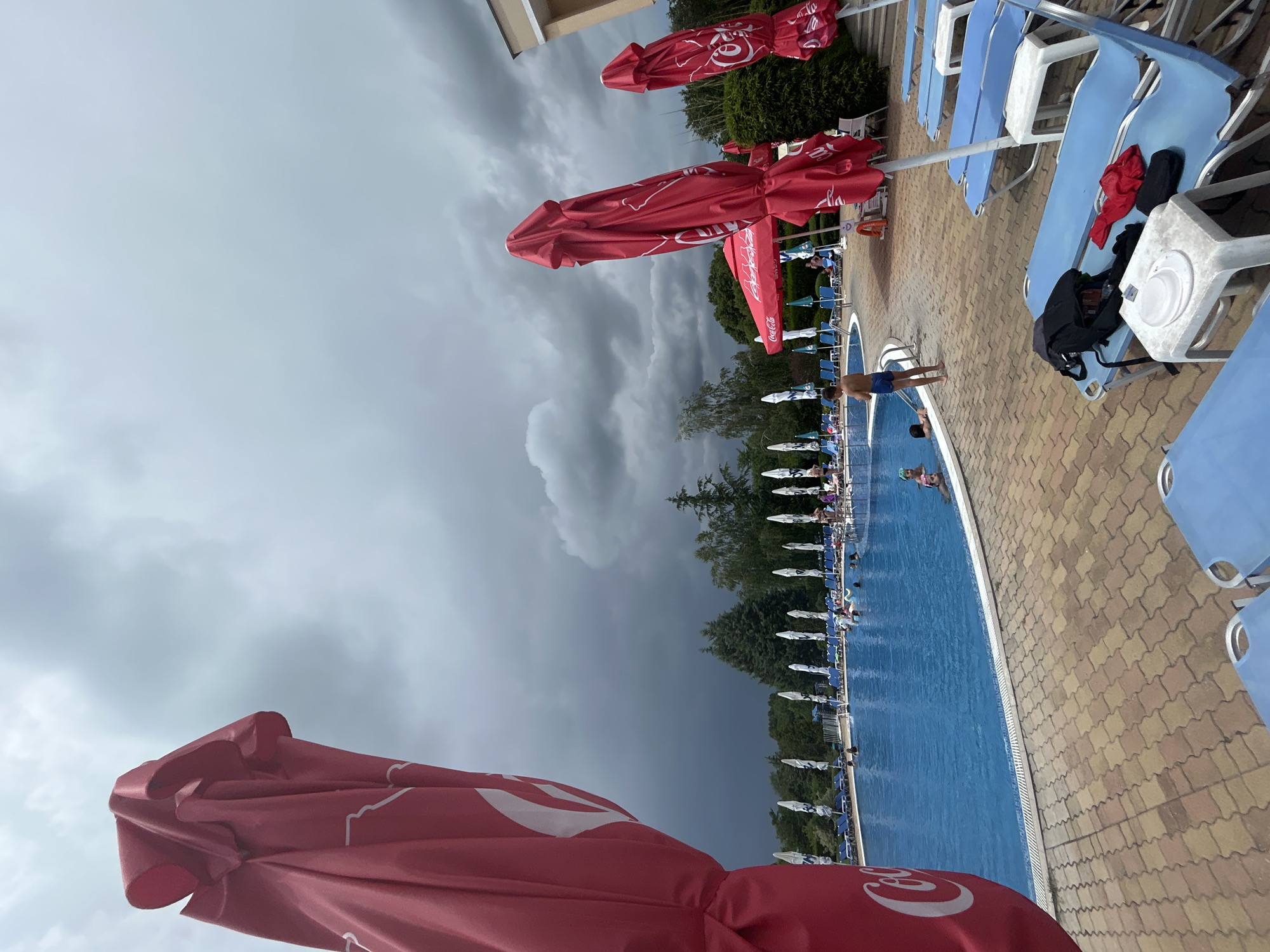 The image shows a crowded outdoor swimming pool area on a cloudy day. In the foreground, to the left, a large, folded red umbrella with a white Coca-Cola logo takes up a significant portion of the frame. Beyond it, a blue swimming pool is filled with people, some of whom are swimming or wading. Several lounge chairs are visible in the middle ground and foreground, some with items like a backpack, a red garment, and a white hat on them. A row of closed white and green umbrellas line the edge of the pool further back. Trees and other foliage are visible in the background, under a dark, overcast sky that suggests impending rain.
