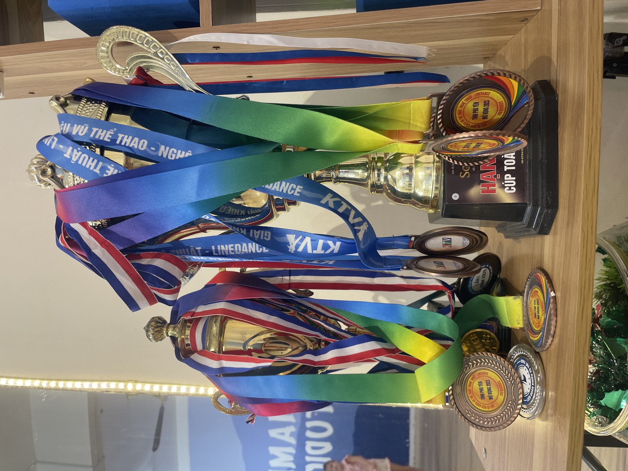 This image showcases a collection of trophies and medals, likely awarded at a competition or event. The trophies are gold in color and have ribbons of various colors (blue, red, white, green, yellow) attached to them. The medals are bronze and silver. Text on the medals mentions "TINH PHU YEN MO RONG 2023," "GIAI KTVX," "LANI NAM 2025," and "CUP TOA." One ribbon reads "XU VU THE THAO-NGHE THUAT." The items are arranged on a wooden shelf. Based on the text present on the trophies and medals, it's likely the setting is associated with a competition in Vietnam, possibly in the city of Bac Nha Trang Ward in Vietnam.
