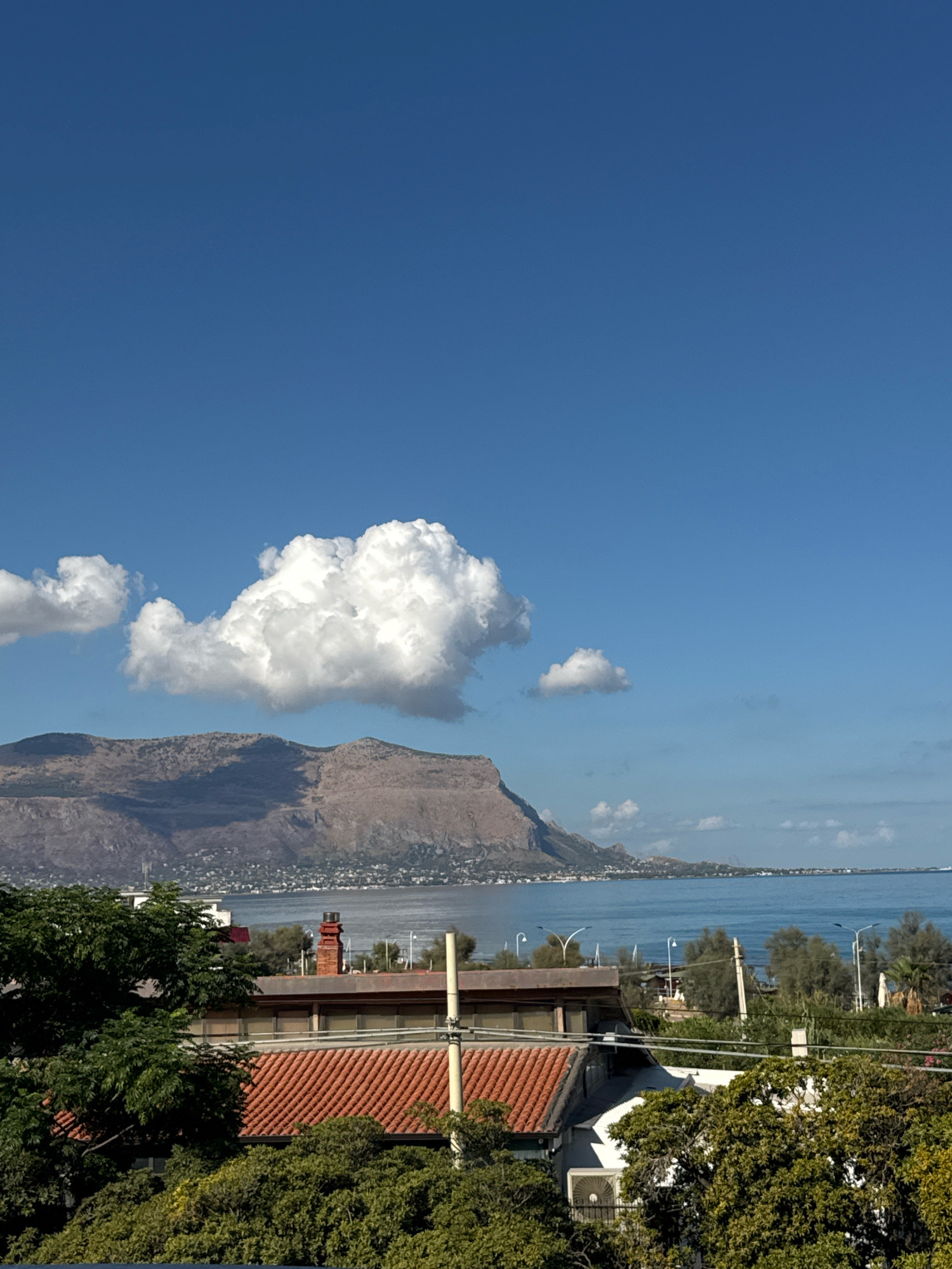 This is a photograph taken in Capaci, Italy, on a clear day with a vibrant blue sky. In the foreground, a portion of a building with a red-tiled roof and a brick chimney is visible, partially obscured by lush green trees. Beyond the building, a calm, blue body of water stretches towards a mountainous coastline in the distance. The mountains appear arid and sparsely vegetated, with a town or settlement dotting the foothills along the water's edge. Several white clouds are scattered across the sky, with a larger, more prominent cloud formation to the left of the frame. Streetlights and some infrastructure are visible along the coastline, suggesting a populated area. The overall scene evokes a peaceful, scenic coastal landscape.