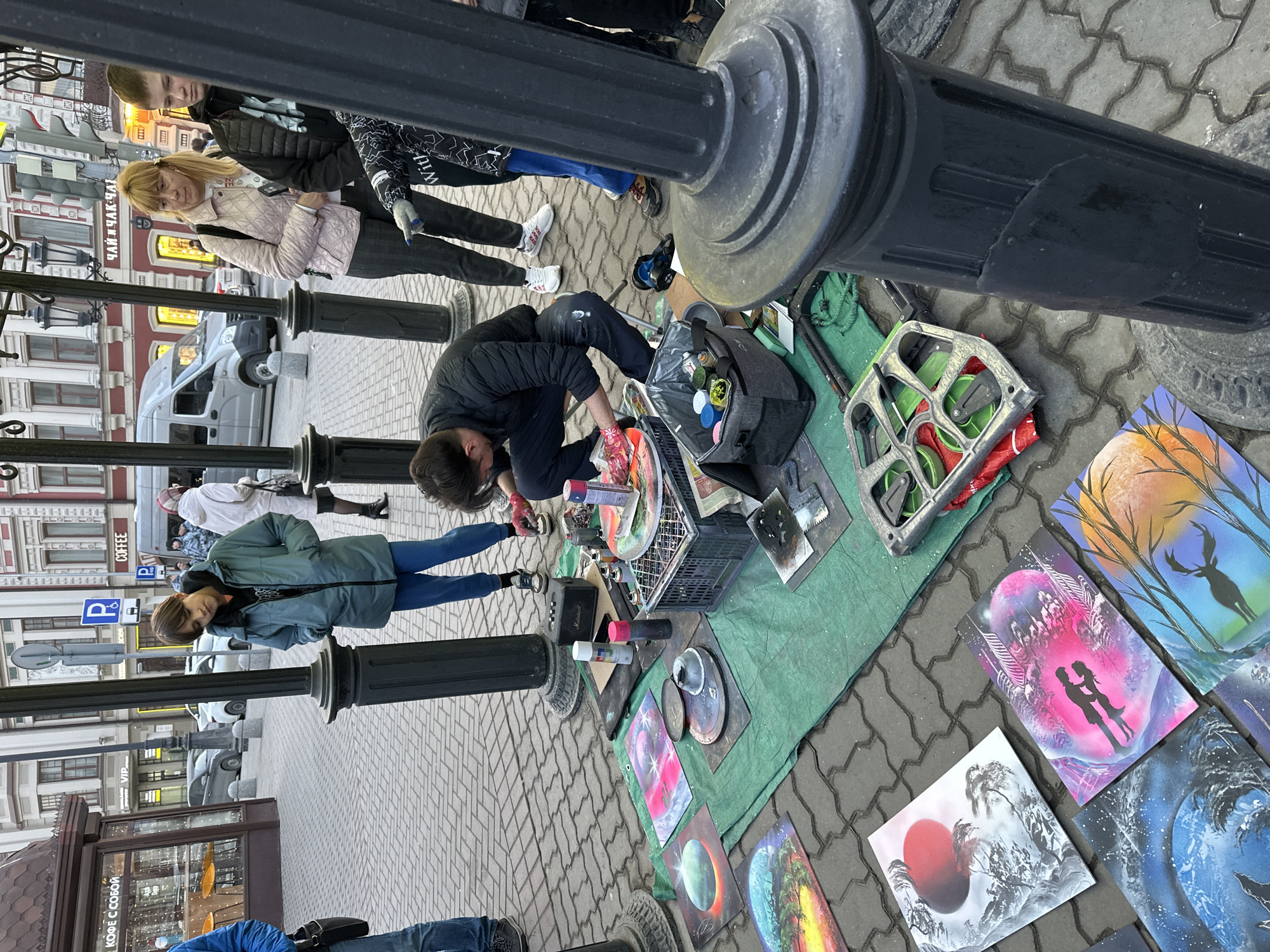 The image depicts a street artist engaged in spray paint art on a cobblestone pedestrian street, identified as Bauman Street in Kazan, Russia, indicated by architectural styles and visible shop signs such as "ЧАЙ И ЧАК-ЧАК" (Tea and Chak-chak) and "КОФЕ С СОБОЙ" (Coffee to go).
In the foreground, a male artist with dark hair, wearing a black puffer jacket and dark trousers, is crouched, actively painting a circular canvas with spray cans while wearing pink gloves. His workspace is set up on a green tarp spread over the cobblestones. The setup includes an array of aerosol spray cans, stencils, a black Marshall portable speaker, various painting tools, and plastic crates containing supplies. Finished pieces of art, primarily depicting celestial bodies, planets, silhouetted figures, and abstract landscapes in vibrant colors, are displayed on the tarp and surrounding pavement.
Several individuals are observing the artist. To the left, a male in a teal jacket and blue pants leans against a black ornamental lamppost, hands in pockets. To the right, a blonde-haired female in a pink quilted jacket, dark pants, and white sneakers stands, looking towards the artist. Beside her, a male youth in a black hooded jacket points with his right hand towards the artist's setup. A black ornamental lamppost occupies the immediate right foreground, partially obscuring the view.
The background reveals a bustling city environment. Multi-story buildings with detailed facades, some featuring red brick and white decorative elements, line the street. Beyond the immediate pedestrian area, a street with parked cars and a light grey van is visible. Street signs, including a blue parking sign, and traffic lights are present. The sky is overcast.