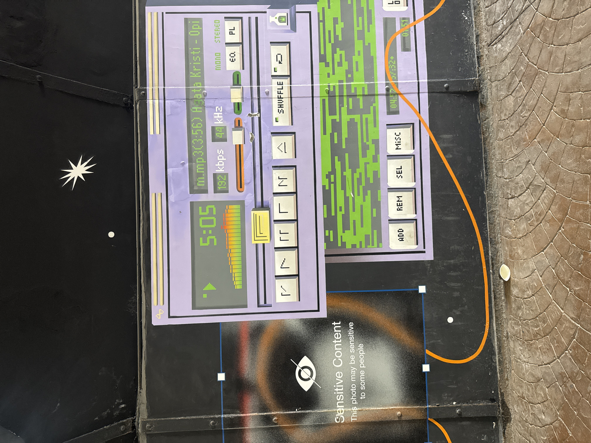 This media file appears to be a photograph of a mural or painted artwork on a dark, textured surface, possibly a wall or a large panel. The artwork itself depicts a stylized representation of a digital audio player or mixer, rendered in shades of purple, green, and yellow.
The dominant subject is the graphic interface of the audio player. It features a digital display showing "5:05" and "04:12 57:32+". There are also labels indicating "m.mp3(3:56) Hata Kristi - Opi", "192 kbps", and "44 kHz", suggesting details about an audio file. Control buttons with labels like "MONO", "STEREO", "EQ", "PL", "SHUFFLE", "ADD", "REM", "SEL", and "MISC" are visible, along with graphical elements resembling sliders and audio level meters. The green sections of the artwork have a pixelated, grid-like design.
To the left of the audio player graphic, there is a white, star-like symbol on the dark background. To the right, the artwork transitions to a textured, stone-like pattern, possibly representing paving stones or a similar surface.
A separate overlay in the image contains text stating "Sensitive Content" and "This photo may be sensitive to some people," along with an icon of an eye with a diagonal line through it. This indicates that the image itself, or its content, has been flagged for potential sensitivity.
The overall scene is static, depicting a piece of street art or mural. There are no people visible in the artwork or the surrounding environment. The lighting suggests it is daytime, but no specific weather conditions are evident. The context provided states the location is Yekaterinburg, Russia, which is a factual detail about the origin of the media file.