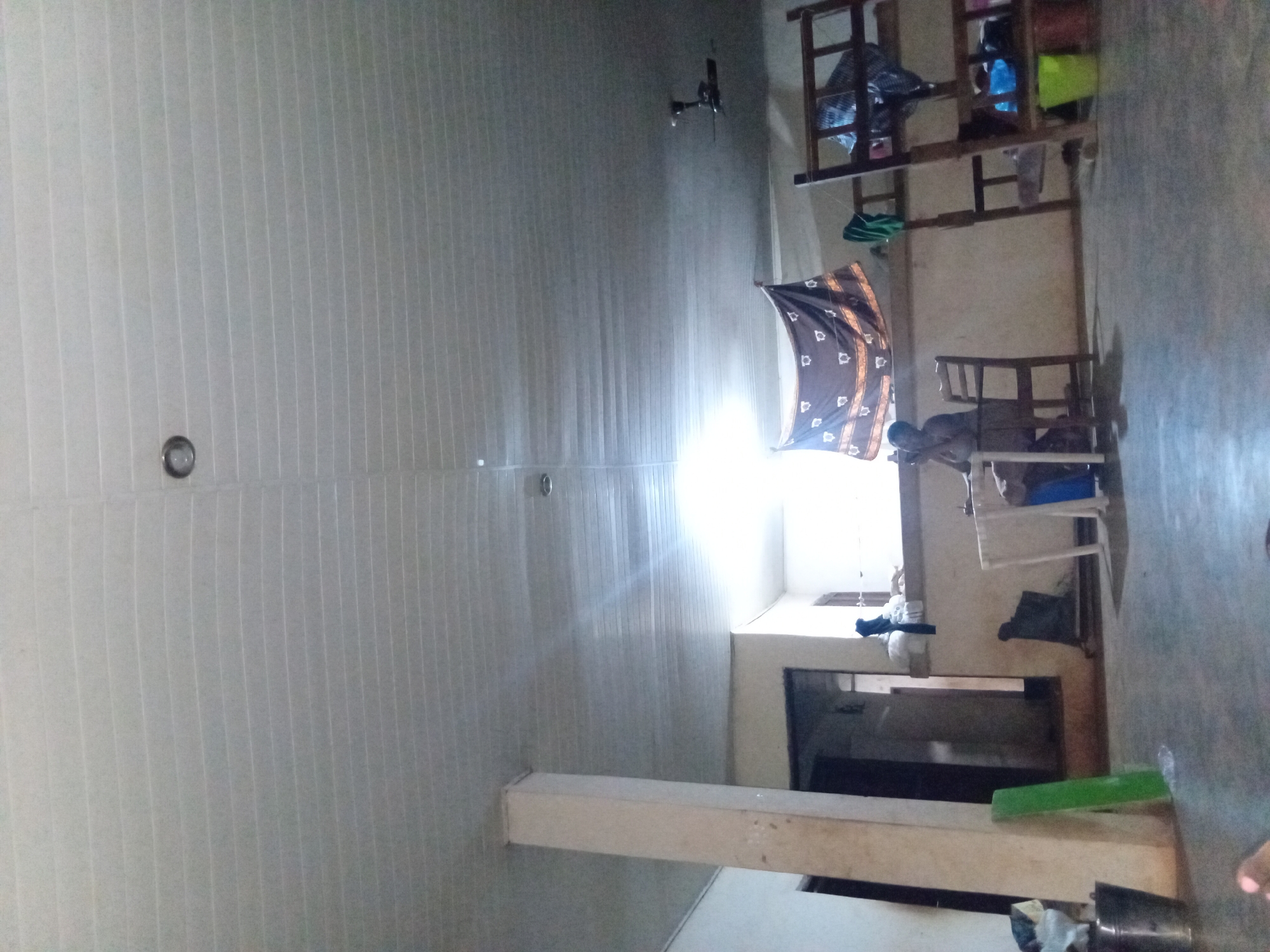 A wide shot captures a dimly lit interior space, possibly a dormitory or communal living area, in Adoma, Nigeria. The ceiling is made of white, grooved panels, with visible fluorescent lights creating a stark illumination. A large, cream-colored pillar stands prominently on the left side of the frame. In the background, a doorway leads into a darker room. A patterned textile, in shades of brown and white, hangs from the wall above a doorway. A person is seated at a white table, facing away from the camera, with a chair beside them. To the right, a wooden bunk bed structure is visible, with items stored on its shelves. The floor is concrete, appearing worn and dusty. In the foreground, a metal bucket rests on the floor. The overall impression is of a functional but somewhat unkempt living space.