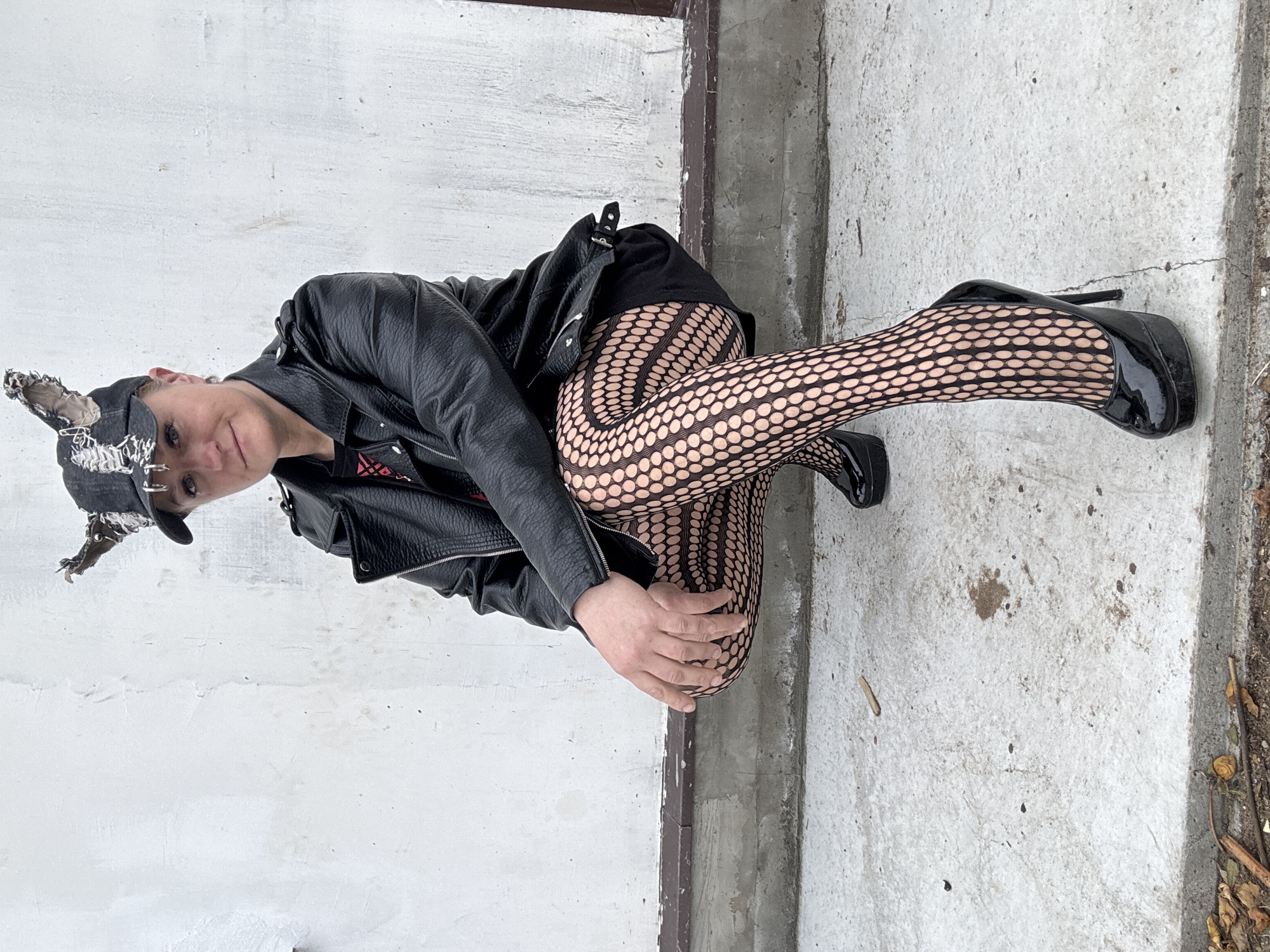An individual wearing a black leather jacket, black tights with a dot pattern, and black high-heeled shoes is crouching in an outdoor environment. The person is also wearing a black cap with some adornments or designs on it. The background consists of a textured white wall on one side and a concrete surface on the other. There is debris along the edge of the concrete. The image's orientation is rotated clockwise. The location is unspecified and cannot be determined from the image.