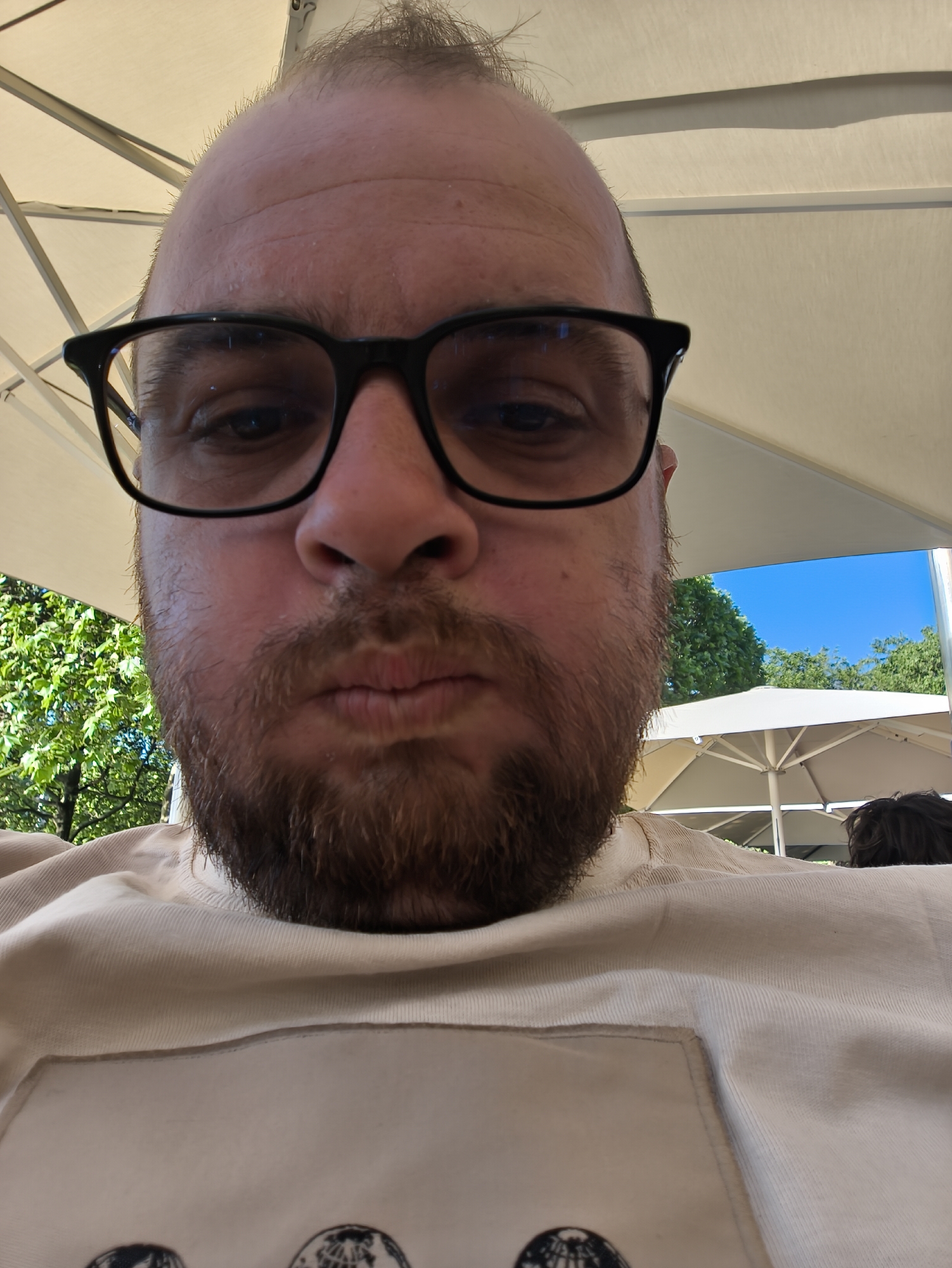 A close-up shot shows a man with a beard and glasses, looking directly at the camera with slightly pursed lips. He is sitting outdoors under a white umbrella, visible in the background along with green trees and a bright blue sky. The man is wearing a light-colored t-shirt. Another person is partially visible behind him. The location is Unknown, Unknown.