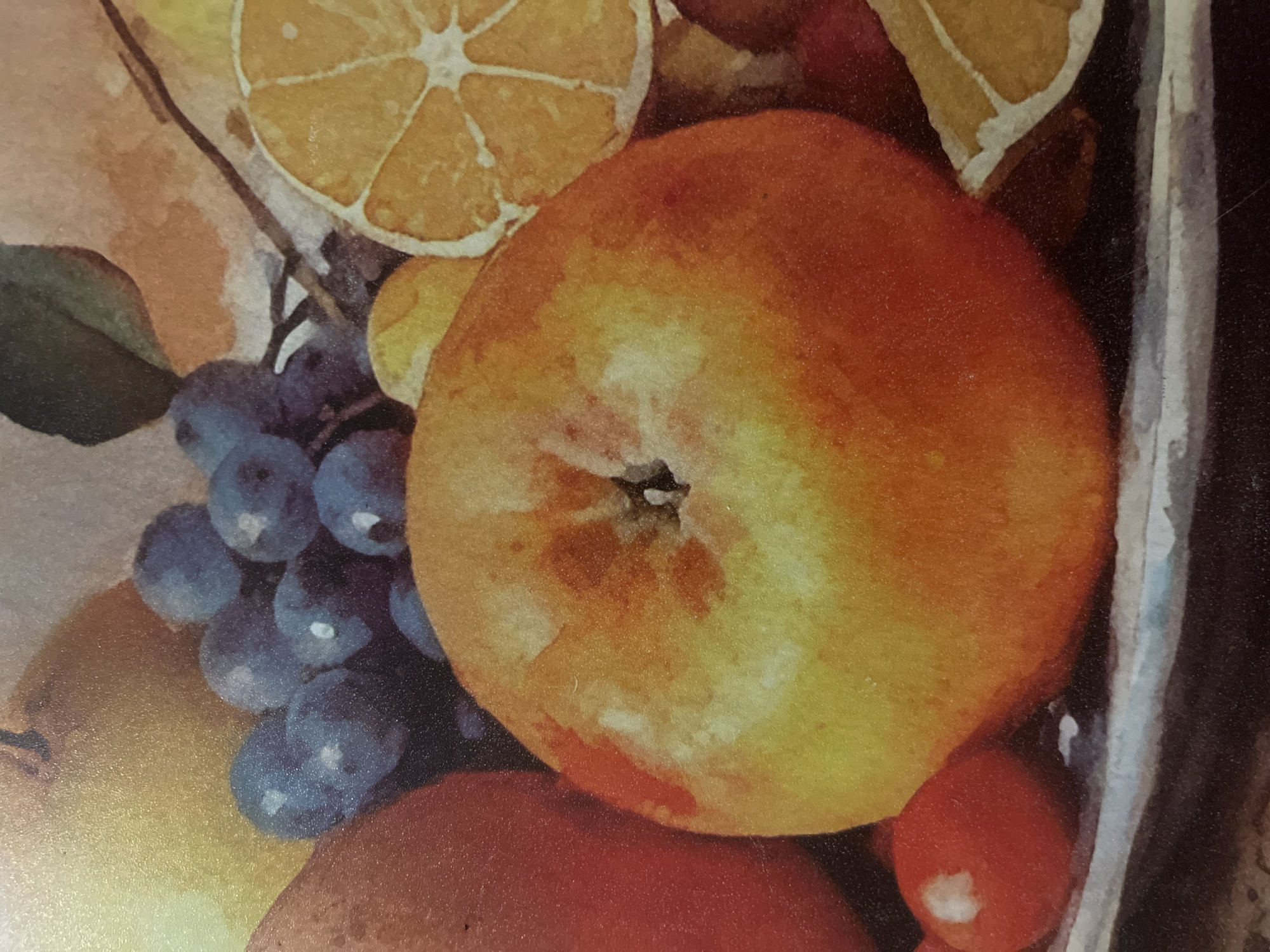 The image is a close-up, high-angle photograph of a still life painting. The painting depicts a variety of fruits, including an apple, grapes, and citrus slices, arranged on a platter. The apple is prominently featured in the foreground, with its skin showing a gradient of yellow and orange. To the left of the apple, a red fruit, possibly a plum or tomato, is visible. Behind the apple, a cluster of dark blue grapes is depicted, along with slices of orange. The background is softly blurred, suggesting a painterly style. The overall impression is of a vibrant and colorful depiction of fresh produce. There is no information in the image that would indicate a location in Minsk, Belarus.