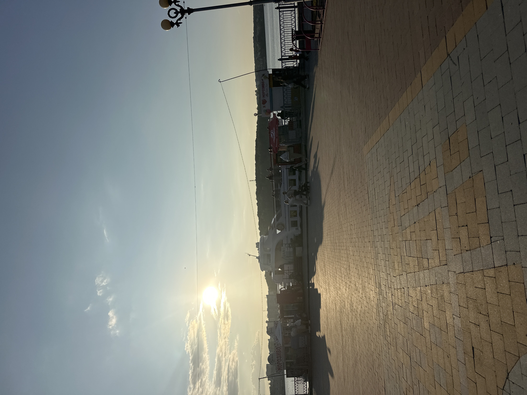 The image depicts a waterfront promenade in Ternopil, Ukraine, bathed in the warm glow of a setting sun. The foreground is dominated by a patterned brick walkway, with a distinct yellow stripe forming a directional arrow. In the middle ground, a riverfront is visible, lined with a decorative fence. Several kiosks offering refreshments, including one with "Морозиво" (ice cream) signage, are present. A white passenger boat is docked alongside the promenade. Several individuals are scattered throughout the scene, some walking, others appearing to be patrons of the kiosks. A lamppost with three globes stands on the right side of the frame. The background features a tree-lined bank across the river and buildings in the distance. The sky is clear with scattered clouds, and the sun is low on the horizon, casting long shadows across the walkway.