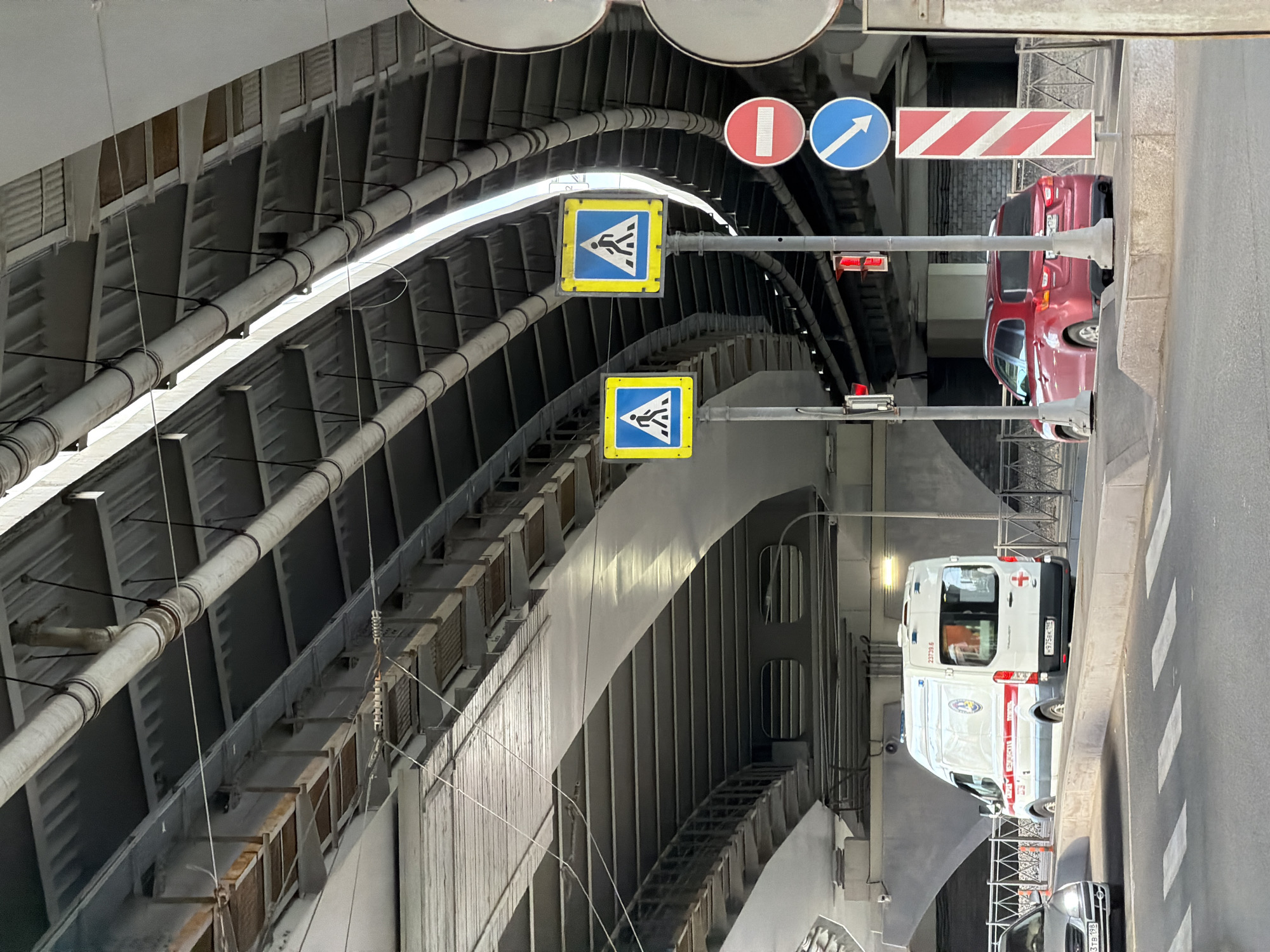 The image depicts a multi-lane road passing through an underpass or tunnel, framed by a robust overhead structure with visible pipes and ventilation systems. The perspective is slightly angled, looking towards the exit or entrance of the underpass.
On the right side of the road, a white ambulance van, possibly a Ford Transit, is parked near a marked pedestrian crossing. The ambulance has a red cross emblem, the text "Медицинская служба" (Medical Service), and a partially visible serial number "23739.5" on its side. Its Russian license plate "H 975 EK 198 RUS" is clearly visible, with "198 RUS" indicating the Saint Petersburg region. Further ahead, a red SUV, likely a Mitsubishi Outlander, is also parked or stopped. Part of another silver car is visible in the foreground.
Several road signs are prominent above the road: two yellow square signs with blue borders indicating a pedestrian crossing, a red circular "No Entry" sign, a blue circular "Direction to Follow" sign (pointing diagonally up-right), and a rectangular red and white striped warning sign, typically used for temporary obstructions or roadworks. A traffic light showing a red signal is also visible. The road itself features white pedestrian crossing stripes and lane markings.
The underpass structure is made of grey metal and concrete, featuring ribbed panels and exposed utility pipes running along the ceiling. Lighting fixtures are integrated into the structure. The scene appears to be set during daytime with bright, possibly overcast, lighting conditions, and no adverse weather is apparent. The visible Russian license plates and medical service text strongly confirm the location as Saint Petersburg, Russia.