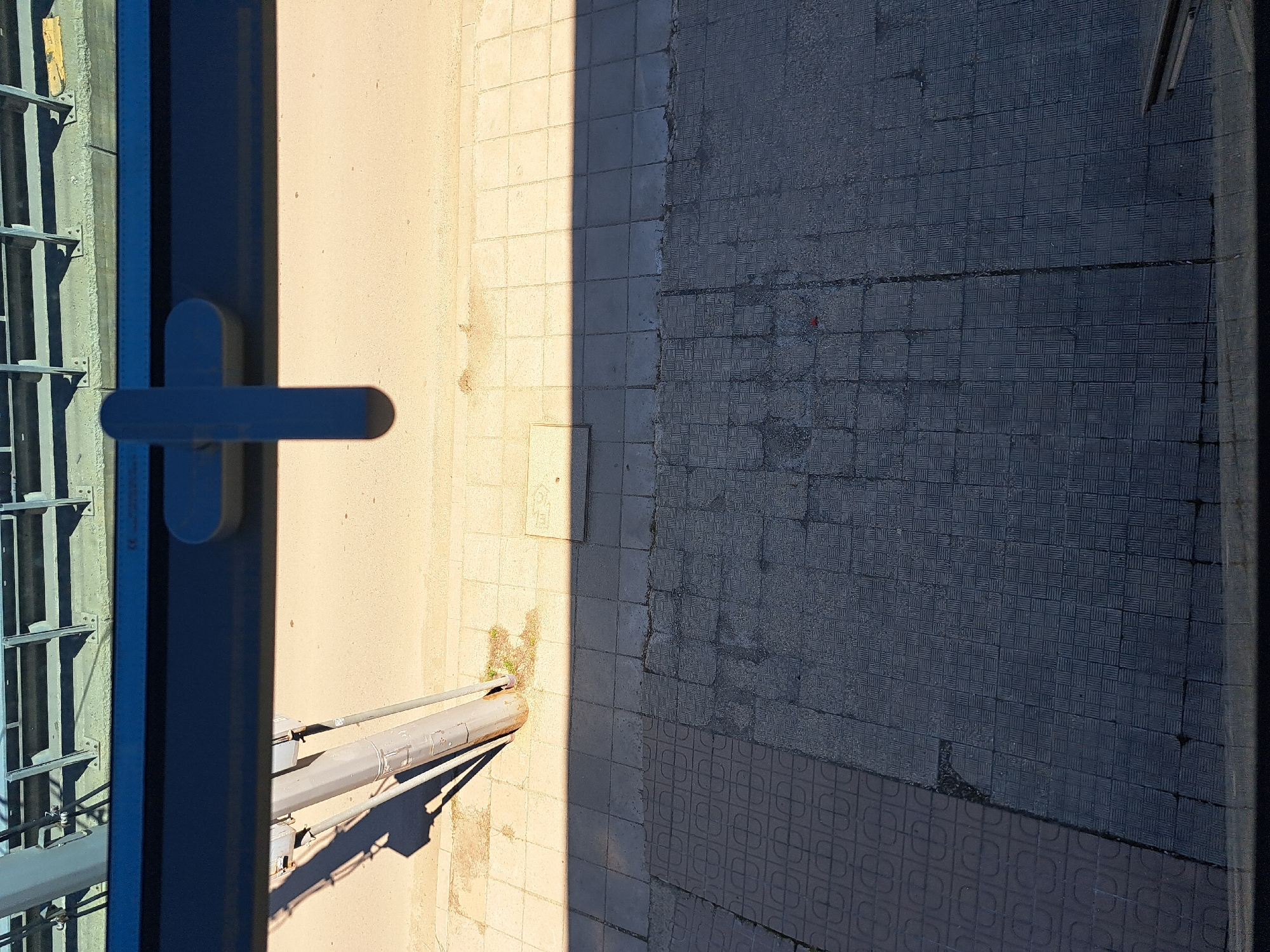 An elevated view from an open window shows a sunlit sidewalk in Sofia, Bulgaria. A thick, blue-painted metal window frame occupies the left side of the frame, featuring a window handle and a metal mesh structure. The sidewalk is divided into two distinct sections: one side is lighter in color and possibly made of smaller, smooth, square tiles, while the other side consists of darker, textured square tiles, some arranged in a decorative pattern. A gray pipe runs across the lighter section of the sidewalk. The scene is mostly devoid of any signs of people.