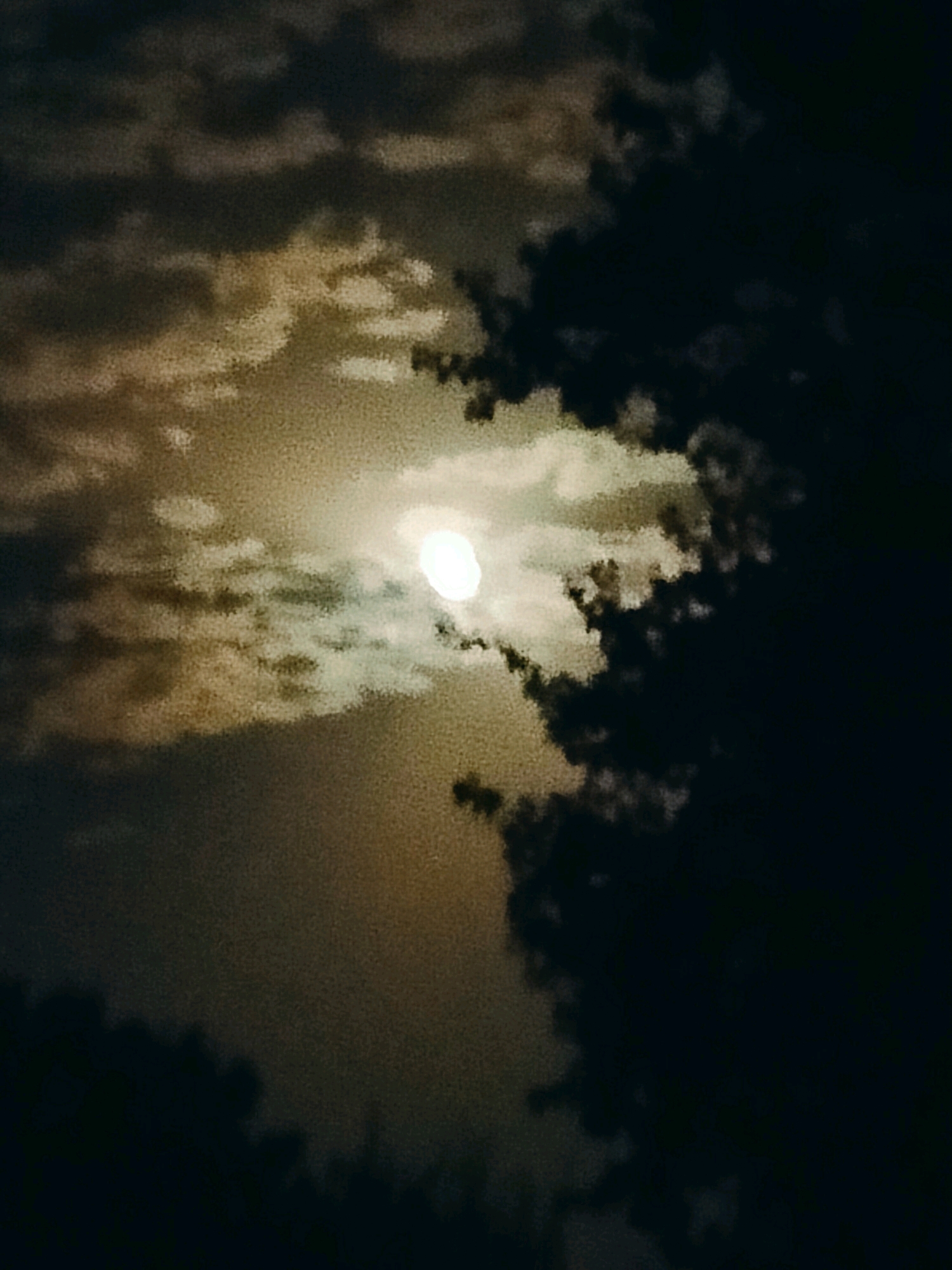 The image shows a cloudy night sky with a bright full moon partially obscured by clouds. The moon is located in the upper center of the frame, emitting a strong, diffused light. Dark, dense clouds dominate the right side of the image, contrasting with lighter, more translucent clouds that surround the moon. The overall atmosphere is moody and somewhat mysterious due to the limited visibility and the dramatic interplay of light and shadow. The scene is not geographically specific to the city of Көлкент, Kazakhstan, as there are no identifiable landmarks or features that indicate this location.