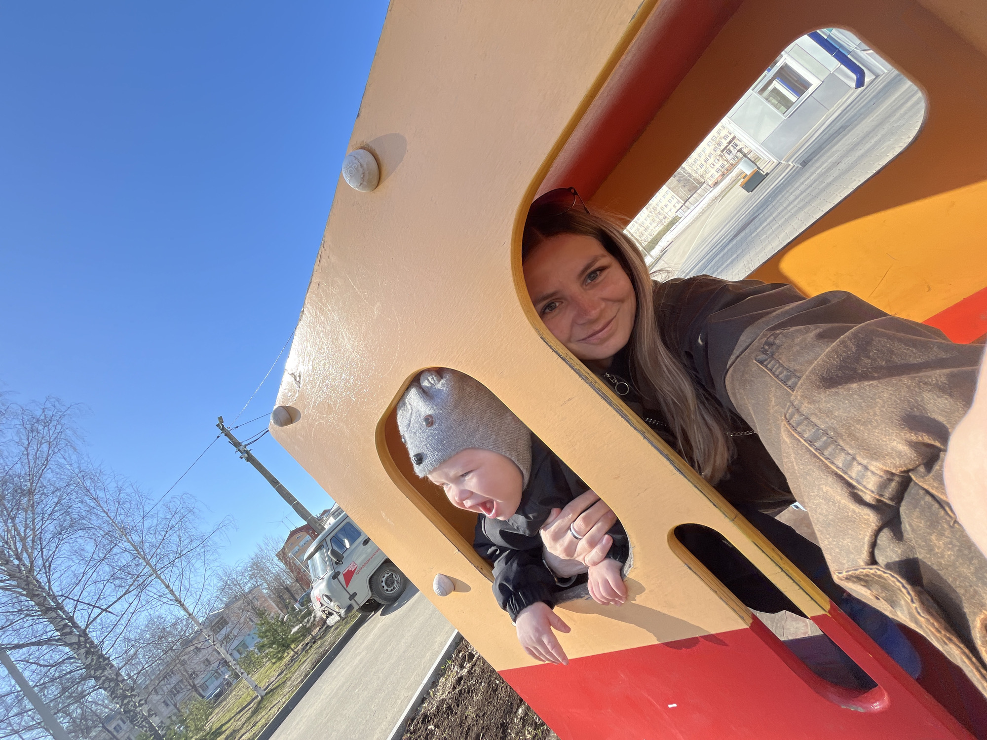 A woman and a child are inside a playground structure, likely a train or a bus, in what appears to be a park in Chaikovsky, Russia. The woman is smiling at the camera, with sunglasses perched on her head. The child, wearing a gray hat, is looking out of a window, mouth open as if speaking or yelling. The structure is yellow and red. Outside the structure, there are trees, buildings, and a white vehicle, possibly a UAZ van. The sky is clear and blue. The location is likely a park or a recreational area.