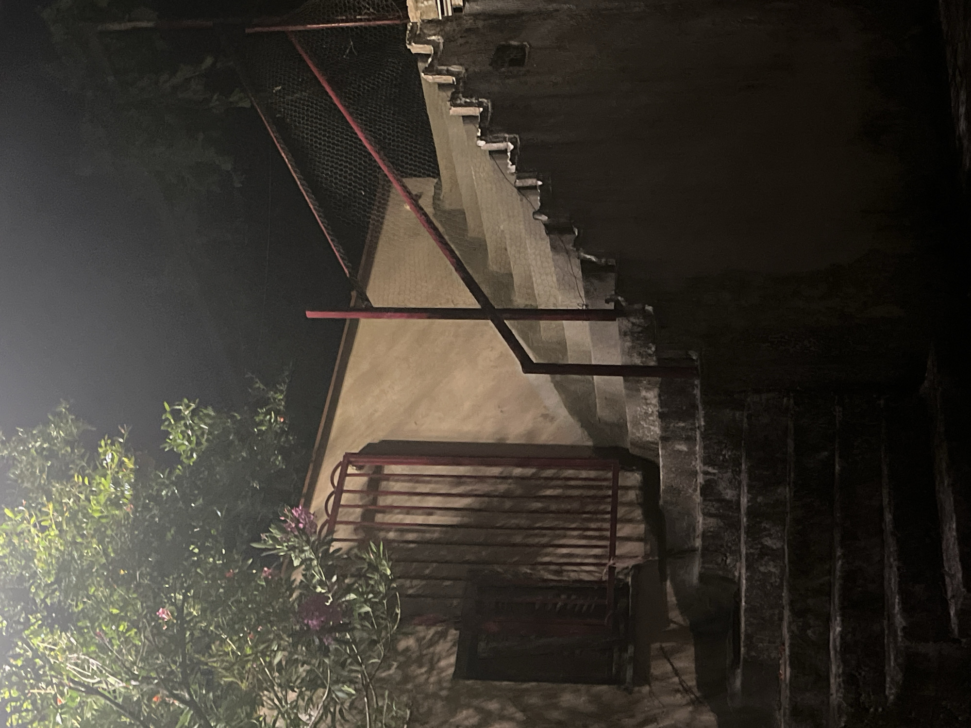 The image appears to be taken at night in an outdoor setting. It features a stone structure that appears to be part of a building with exposed walls and staircases. A metal railing is visible, painted red. Mesh metal hangs over part of the structure. Vegetation is present in the left side of the frame, lit by a light source. The scene is dimly lit. The location can't be determined with certainty, nor can actions or interactions be discerned from this image.