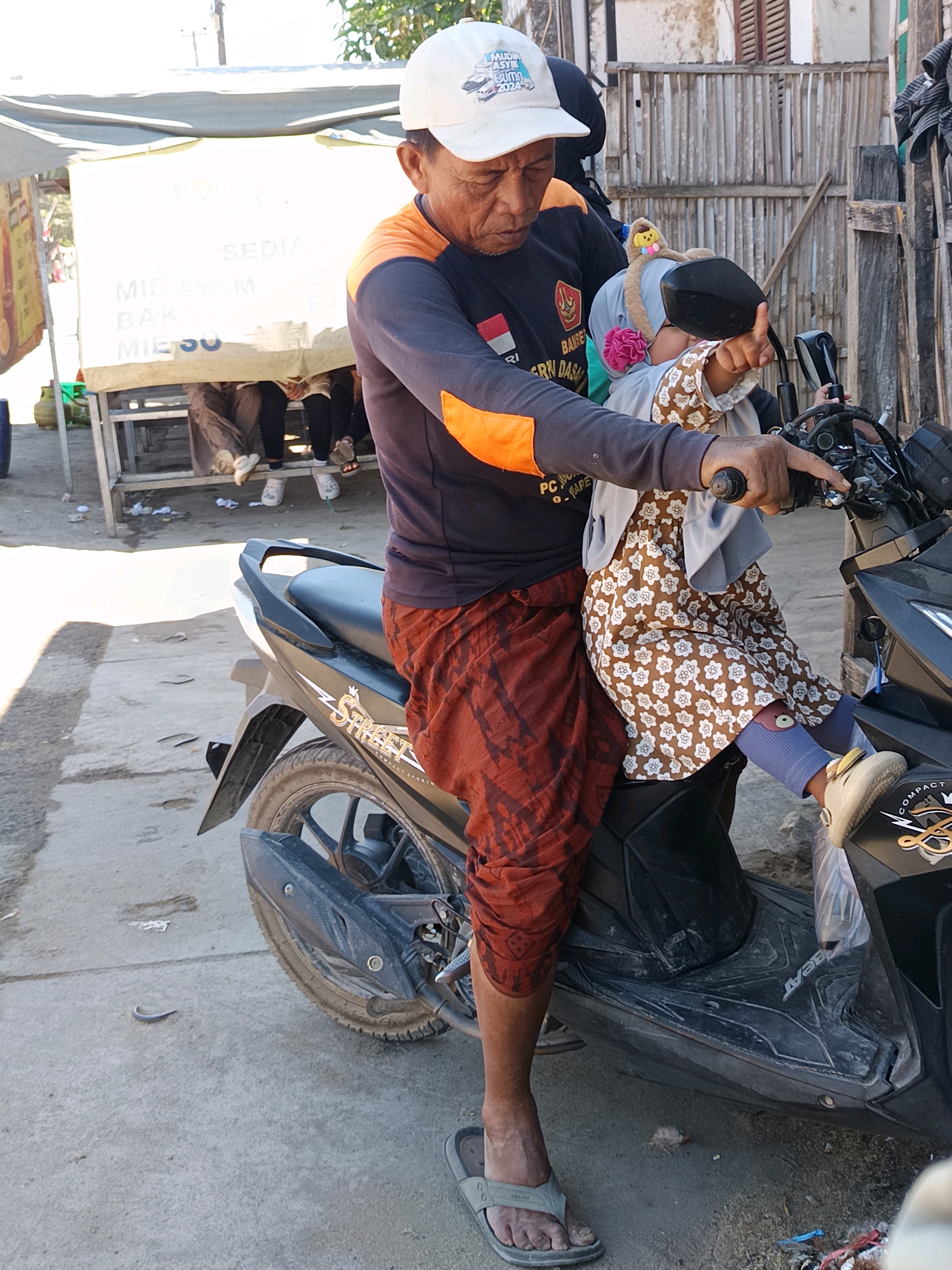 An elderly Indonesian man wearing a white cap and a dark long-sleeved shirt with orange accents is seated on a dark-colored scooter, holding a young child in his lap. The man has his right hand on the scooter's handlebar and appears to be preparing to ride. The child, wearing a light-colored headscarf with a flower detail and a patterned dress, is looking towards the man. They are positioned in front of a white stall with text that includes "MIE AYAM" and "BAKSO," indicating food items. Several people are visible sitting behind the stall, some wearing masks. The environment suggests an outdoor setting, possibly a marketplace, with a wooden fence in the background. The ground is concrete, and there are various debris and discarded items scattered around. The scene takes place in Blega, Indonesia, as indicated by the text on the white stall.