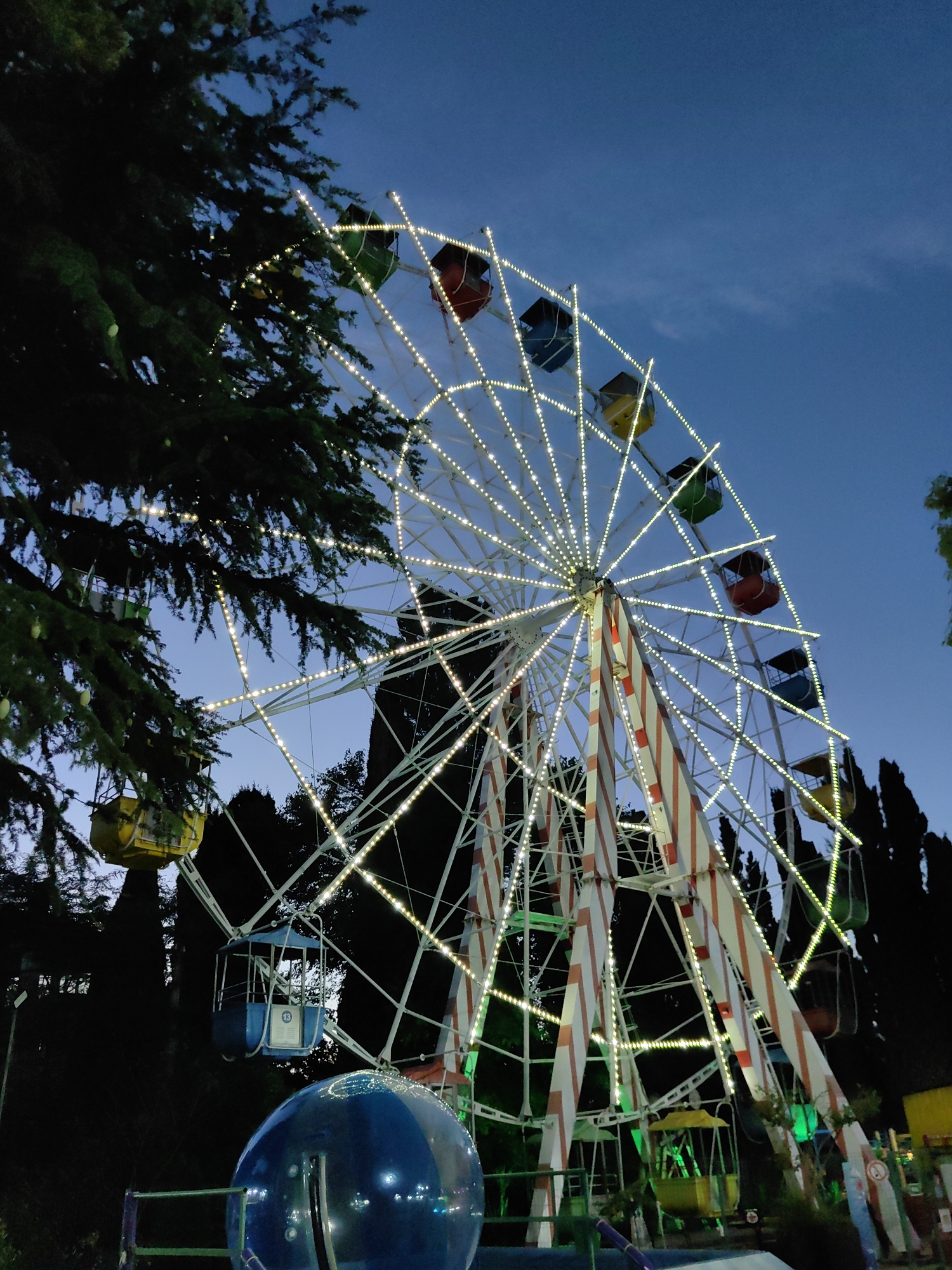 This is a medium-shot, eye-level, outdoor photograph taken at dusk in the city of Sochi, Russia. The primary subject is a Ferris wheel, illuminated by numerous small, white lights. The Ferris wheel has a white frame with red and white striped supports. It is adorned with colorful gondolas, including yellow, red, green, and blue ones. A large evergreen tree partially obscures the left side of the Ferris wheel. In the foreground, a large, transparent blue inflatable structure is visible, partially obscuring the base of the Ferris wheel. The background consists of dark silhouettes of trees and a clear, twilight sky. The scene appears to be an amusement park or a similar recreational area.