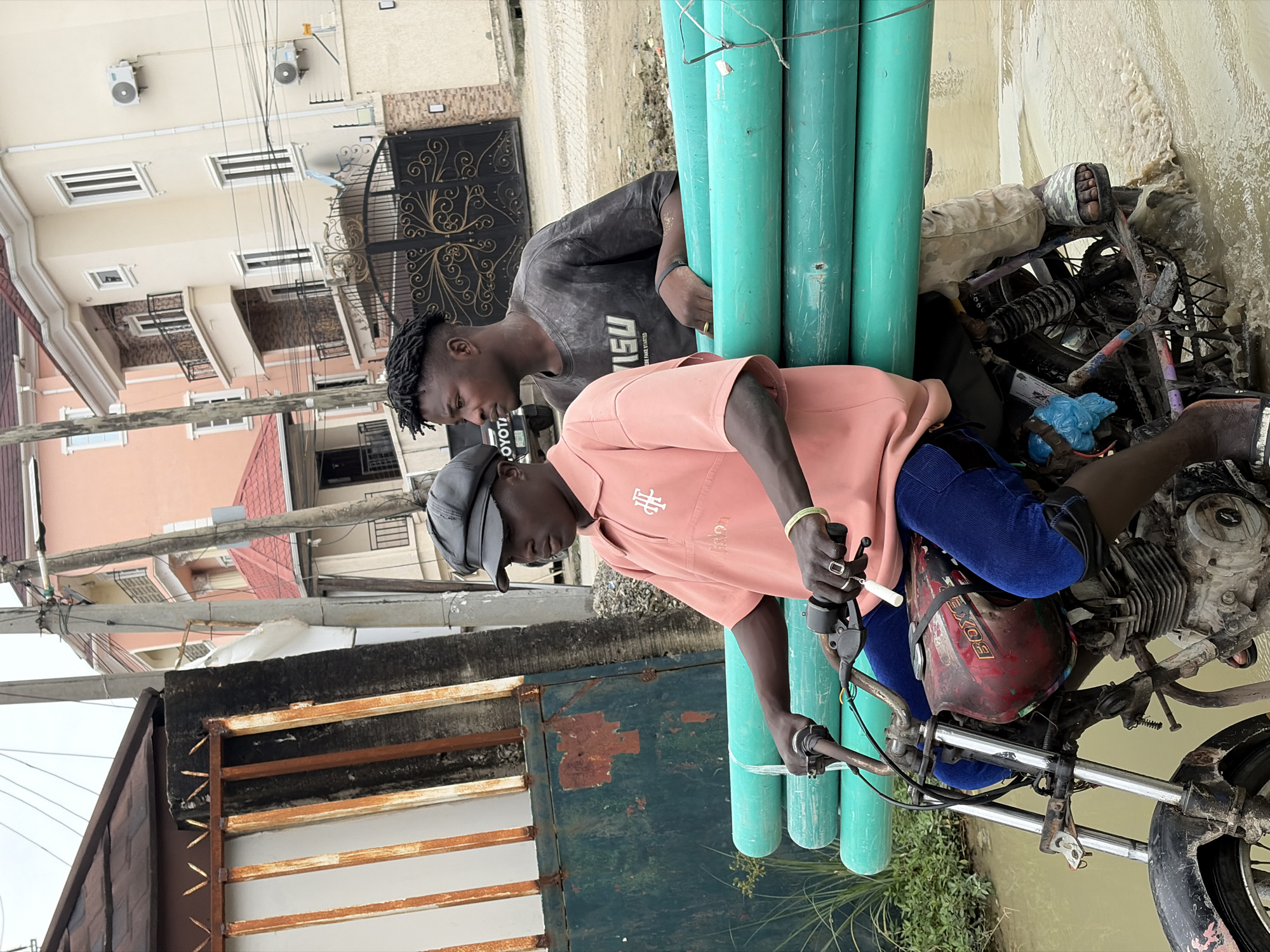 In Igboefon, Nigeria, two young men are riding a motorcycle through muddy water. The driver, wearing a pink polo shirt and a black cap, steers the bike which carries a stack of large green pipes. Behind him, a passenger in a dark grey t-shirt looks to the side. The motorcycle is old and worn, with visible rust and mud. The background shows modern houses with balconies and an ornate gate, contrasting with the rougher foreground. The cloudy sky suggests an overcast day.