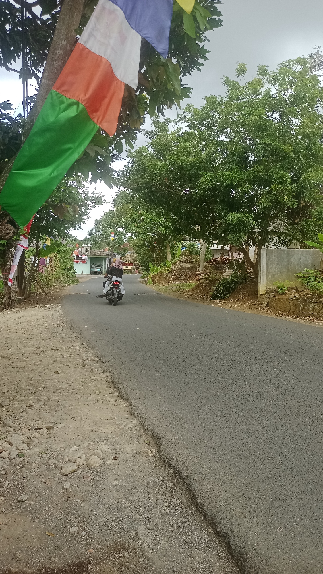 The image depicts a street scene in the city of Cibeber, Indonesia. In the foreground, on the left, is a dirt shoulder with scattered rocks and debris. The asphalt road curves to the right and recedes into the distance. A person is riding a motorcycle away from the camera on the road. Behind the motorcycle, the road is lined with trees and some buildings. In the upper left corner, colorful flags are strung on a tree. The sky is overcast.