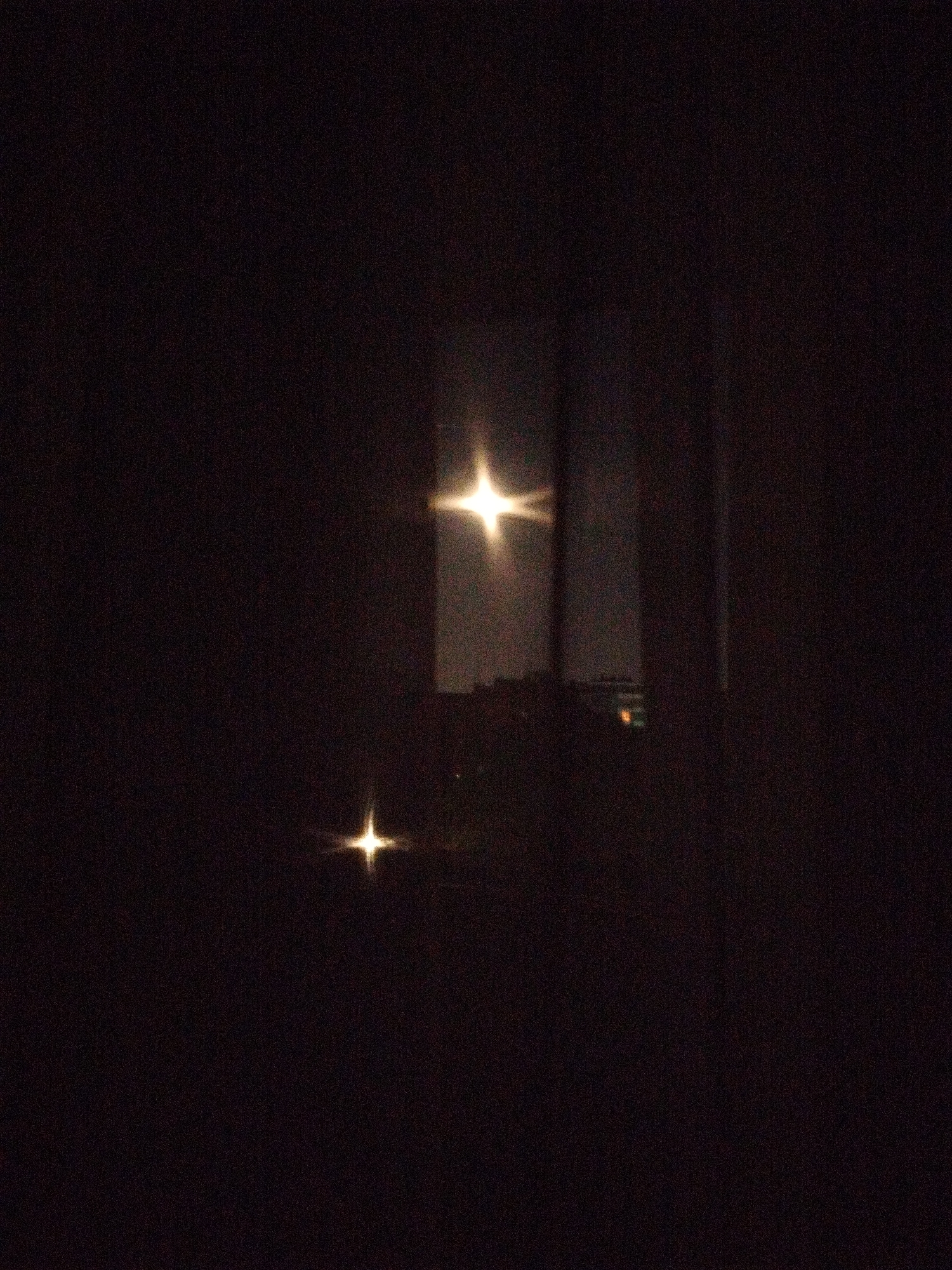 The image is a very dark nighttime scene, possibly taken from indoors looking out a window. Two bright light sources are visible outside, one significantly brighter than the other. The brighter light is higher in the frame, and both lights exhibit a starburst effect. The outlines of buildings and possibly other structures are faintly discernible in the background. Based on the window frame, the scene appears to be viewed through a window. Given the presence of buildings and lights at night, this scene is likely within a city environment. While a specific location in Sofia, Bulgaria cannot be determined with absolute certainty, the presence of structures and night lighting is consistent with an urban context, and the window is a typical urban architecture.