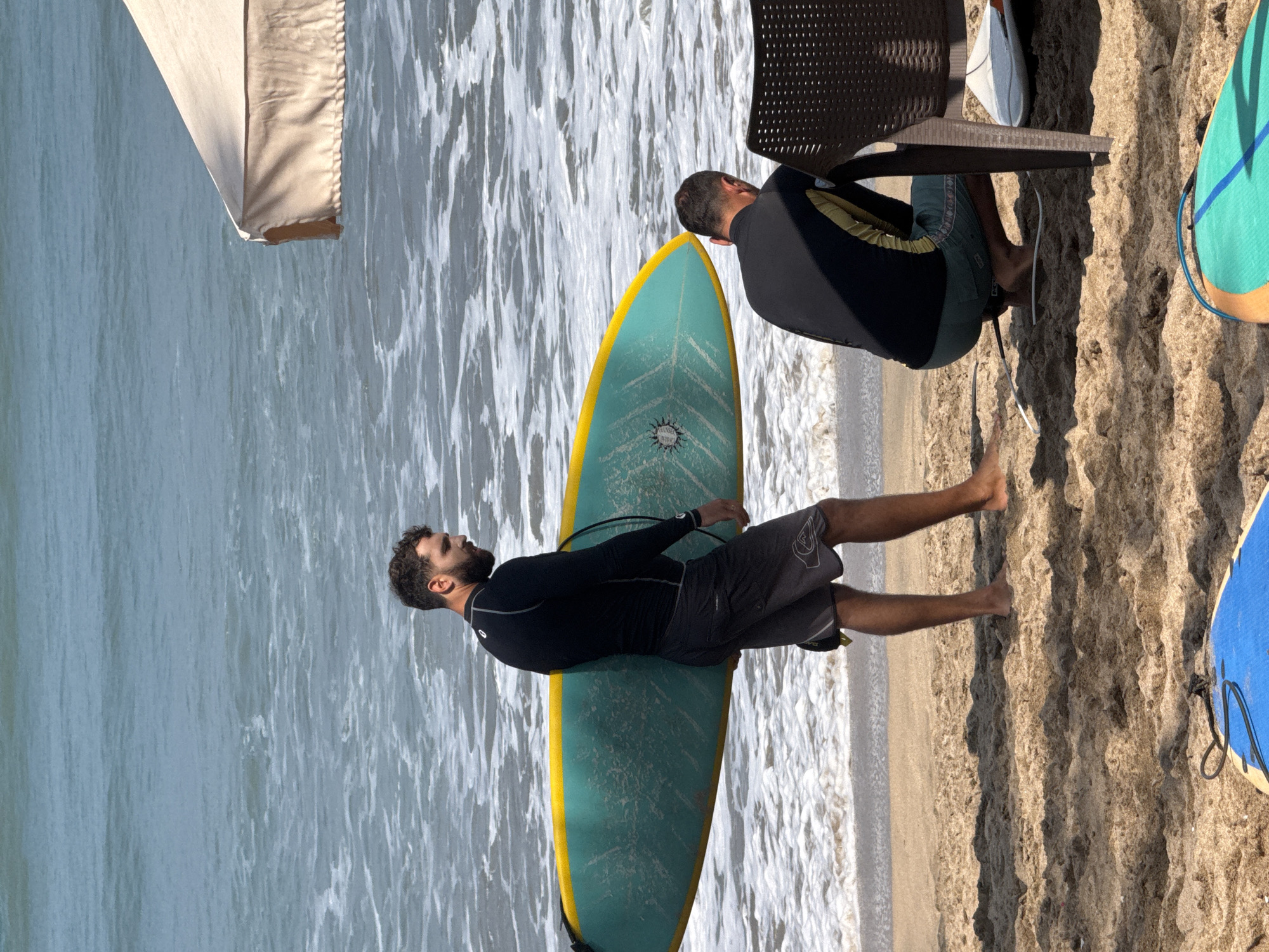 Two men are on a beach in Kuta, Indonesia, near the ocean. One man, wearing a black long-sleeved rash guard and dark shorts, carries a teal surfboard with a yellow edge, walking near the water's edge. The other man is crouched down, facing away from the camera, wearing a black and yellow long-sleeved rash guard and green shorts, next to a brown plastic chair. The ocean has small waves with white foam. A light-colored umbrella is partially visible in the upper right corner. Two other surfboards are in the foreground, one blue and one teal with orange and blue stripes.
