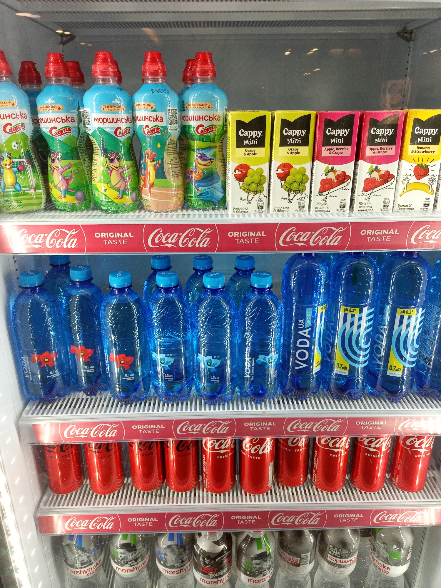 This is a medium shot, eye-level, indoor, waist-up photograph of a commercial refrigerator filled with beverages. The refrigerator has multiple shelves, each displaying a different brand and type of drink. The top shelf contains several bottles of "МОРШИНСЬКА" brand fruit juice, characterized by blue caps and colorful cartoon labels featuring dragons. To the right of these bottles are small carton drinks labeled "Cappy Mini" in various flavors like "Grape & Apple" and "Apple, Berries & Grape." The middle shelf is dominated by bottles of "VODA UA SPORT" brand water, distinguished by their bright blue color, blue caps, and poppy flower labels on some. Below this, a shelf is lined with red cans, likely Coca-Cola, some of which are partially visible and have the "Coca-Cola Original Taste" branding. The bottom shelf shows more "МОРШИНСЬКА" branded bottled water, featuring labels with black and white graphics of sports-related figures. The interior of the refrigerator is brightly lit, and the overall environment suggests a retail setting like a supermarket or convenience store. There are no people visible in the image, and no discernible exterior elements that would indicate a specific city or location.