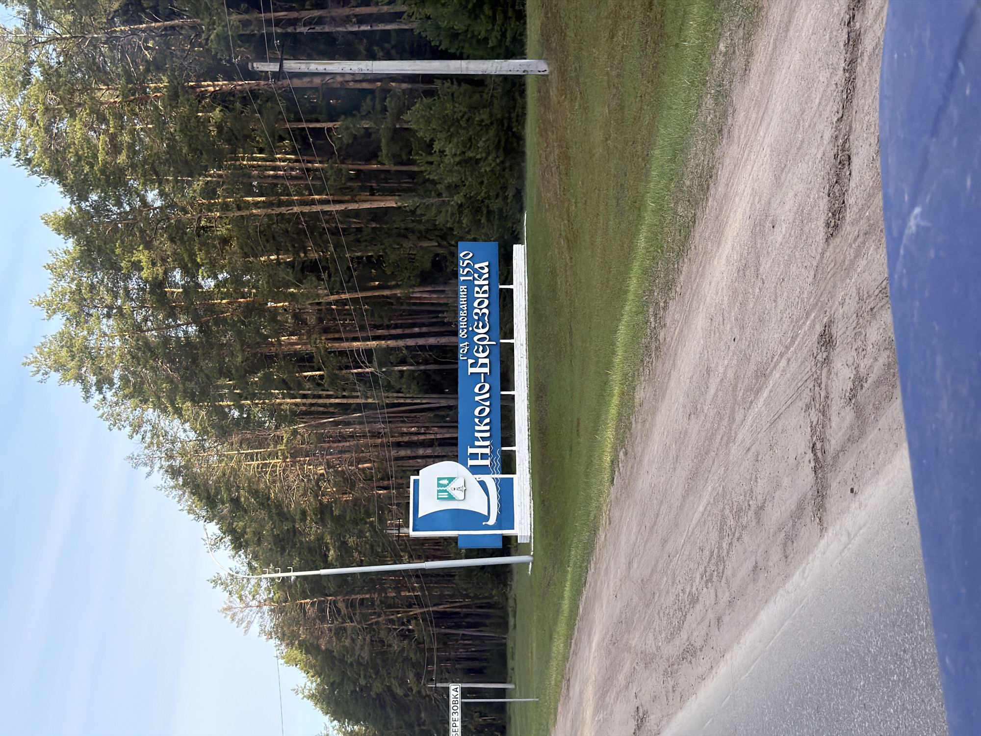 The image displays a roadside scene, depicting an entrance sign to a settlement, set against a backdrop of a dense pine forest under a clear blue sky. The photograph is taken during daytime, with ample natural light indicating sunny weather.
The primary subject is a prominent blue and white road sign, positioned to the right of a dirt or gravel road that features tire tracks. The sign bears text in Cyrillic. At the top, it reads "ГОД ОСНОВАНИЯ 1550" (Year of foundation 1550). Below this, the name of the settlement is clearly visible: "НИКОЛО-БЕРЕЗОВКА" (Nikolo-Beryozovka). To the left of the name, a distinctive crest or logo is displayed, featuring a stylized white boat or ship with a smaller emblem inside, depicting what appears to be a forest and a structure, possibly a church or building. Beneath the main sign, a smaller white sign is partially visible, showing the Cyrillillic word "БЕРЕЗОВКА".
To the left of the road, behind the sign, a thick forest of tall, slender pine trees dominates the landscape, extending across the entire left side of the frame. Power lines and utility poles are visible running through the forest line. The road itself is unpaved, with grass growing on its immediate left edge. On the far right edge of the image, a sliver of a blue object, possibly part of a vehicle, is visible, suggesting the photo was taken from a car or similar transport.
No people are visible, and there is no specific activity or event taking place, beyond the static capture of the scene. The image provides a clear sense of approaching a historical settlement in a rural, forested area, consistent with the context of Nikolo-Beryozovka, Russia. The atmosphere is calm and uneventful.