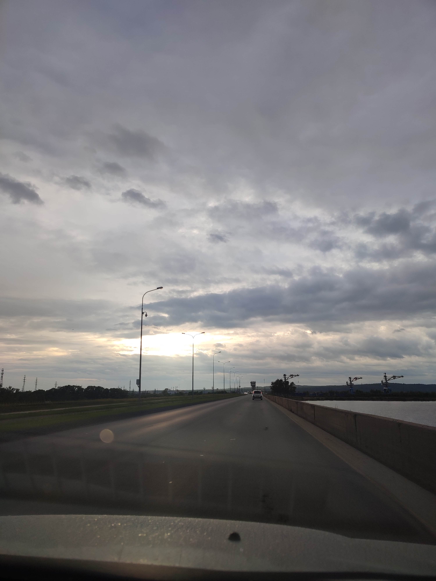 The image captures a first-person perspective from inside a vehicle traveling forward on a multi-lane asphalt road or bridge in the city of Unknown, Unknown. The sky is predominantly overcast with varying shades of grey clouds, and a bright, hazy band of sunlight penetrates the clouds near the horizon on the left, suggesting either sunrise or sunset.
Along the left side of the road, numerous streetlights are visible, extending into the distance. Further left, power lines and associated towers are discernible against the distant landscape. A light-colored sedan is observed traveling ahead in the same direction on the road.
To the right of the road, a substantial concrete barrier separates the thoroughfare from a wide body of water, likely a river or lake. On the far bank of this waterway, several large, dark industrial cranes are prominent, positioned against a backdrop of distant hills or landforms.
The immediate foreground reveals a portion of the vehicle's dark-colored dashboard and windshield, displaying a distinct circular lens flare or reflection approximately in the lower-middle section, along with minor dust particles. No individuals are explicitly visible.