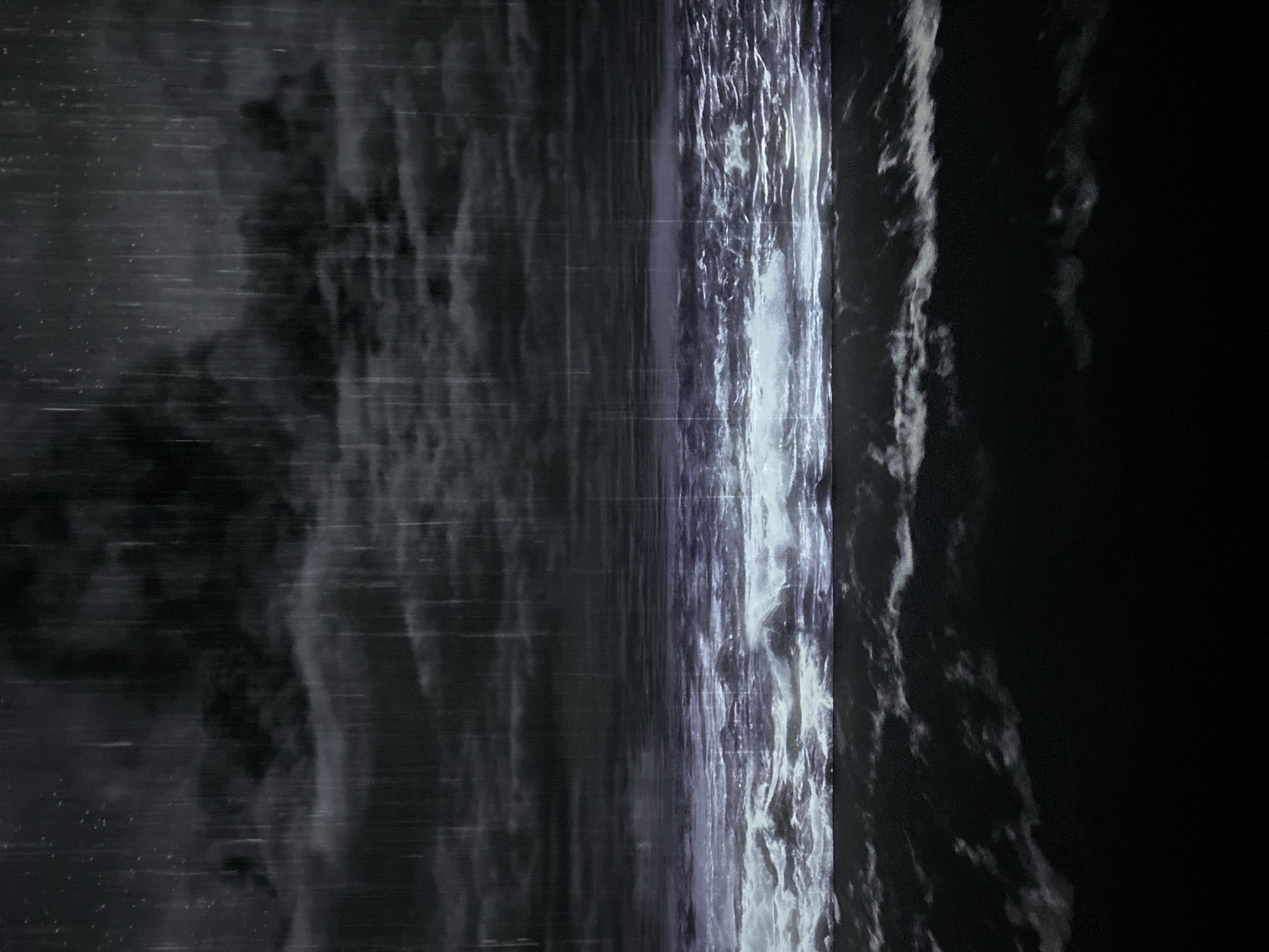 The image depicts a stormy scene at sea, possibly in Pattaya City, Thailand. Heavy rain falls from dark, ominous clouds, creating streaks of light against the dark sky. The choppy ocean waves are illuminated by an unseen light source, giving them a turbulent, almost ethereal appearance. The lower portion of the image is dominated by the dark, foreboding sea. There are no people or man-made structures visible in the scene, emphasizing the raw power of nature.