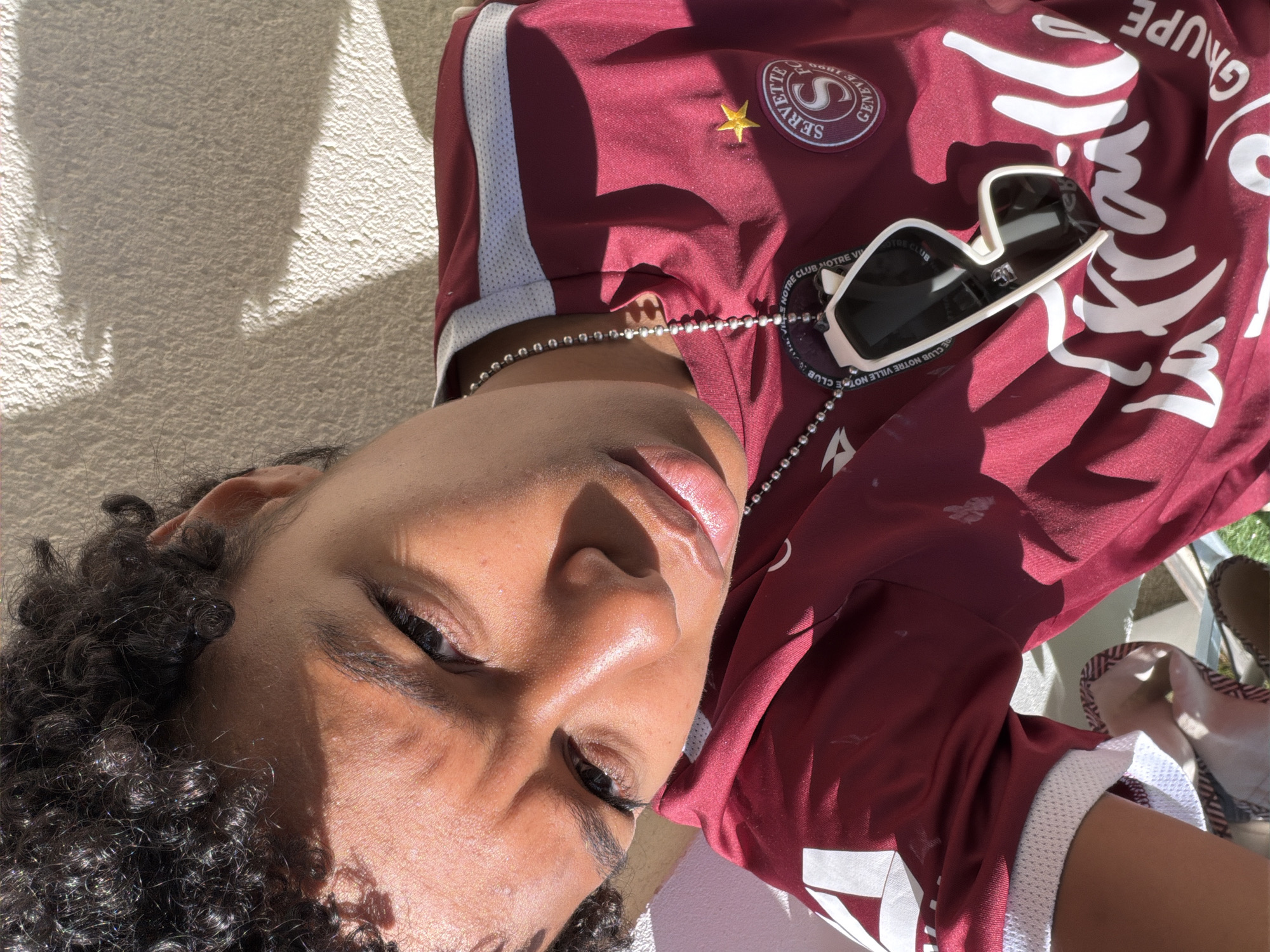 The image shows a person with dark curly hair, brown skin, and dark eyes. They are wearing a maroon jersey with white accents and a logo on the chest with a gold star above it. A silver ball-chain necklace is around their neck, and a pair of white-rimmed sunglasses rests on the jersey. The person is lying on what appears to be a textured concrete surface. The lighting indicates it is daytime and the sun is shining brightly. The writing on the jersey includes the words "CLUB NOTRE VILLE," implying a location or affiliation with a city in France, possibly Angers, France.