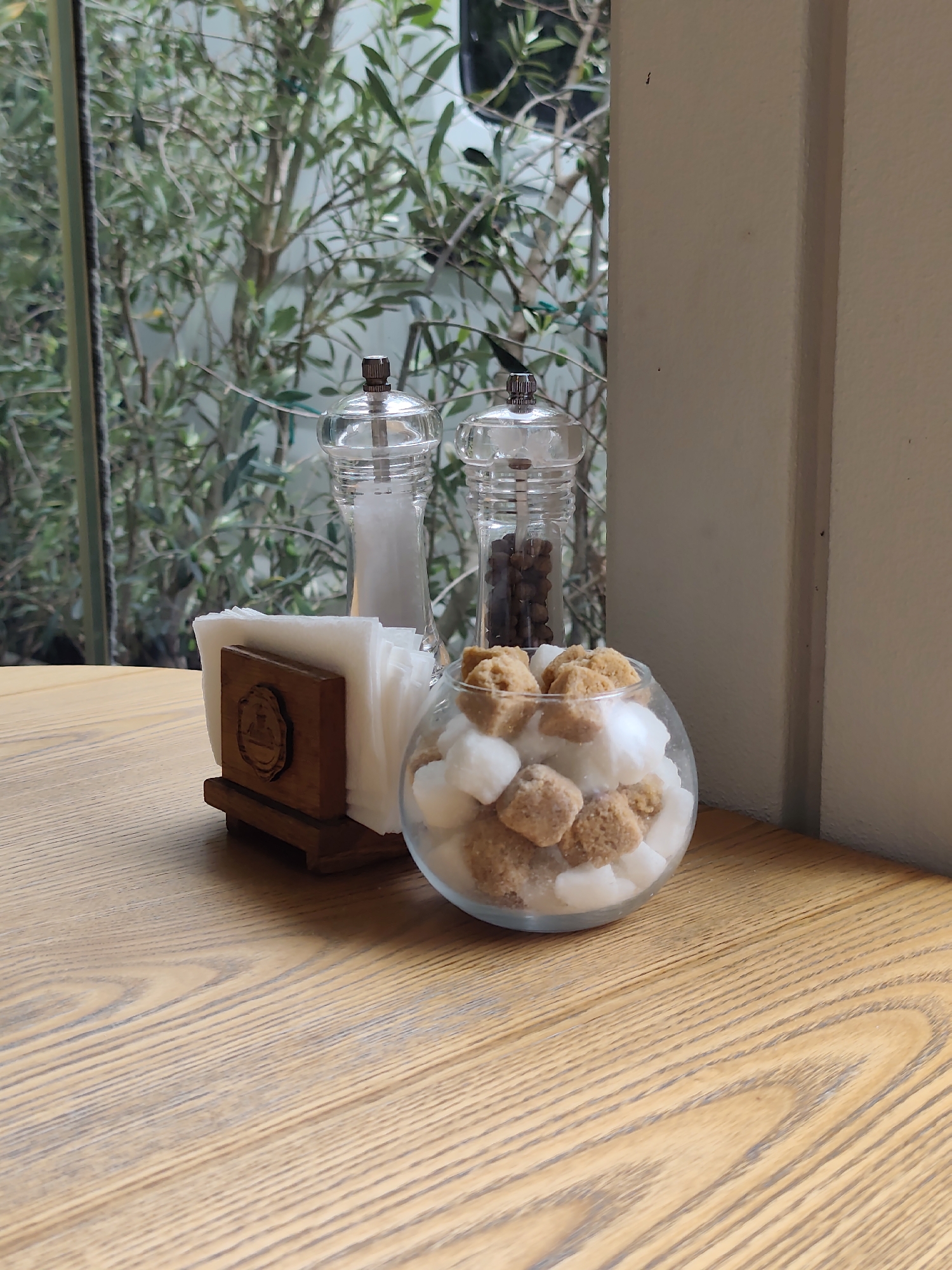 On a wooden table, a clear glass bowl filled with white and brown sugar cubes sits next to a wooden napkin holder containing white paper napkins. Behind the napkin holder are two clear glass shakers, one filled with white salt and the other with black peppercorns. Through a window behind the table, green foliage of trees is visible, suggesting an outdoor or semi-outdoor setting. The scene appears to be a tabletop setup at a cafe or restaurant in Sochi, Russia.