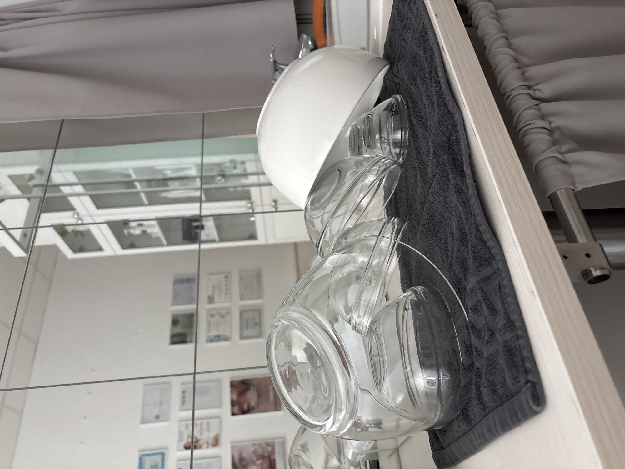 The image displays a stack of clear glass bowls and mugs, topped by a white ceramic bowl, placed on a dark gray towel. The objects are situated near a mirror which reflects a wall decorated with pictures and certificates. A gray curtain is also visible in the background. The location could be Romanovka, Russia, based on the prompt.