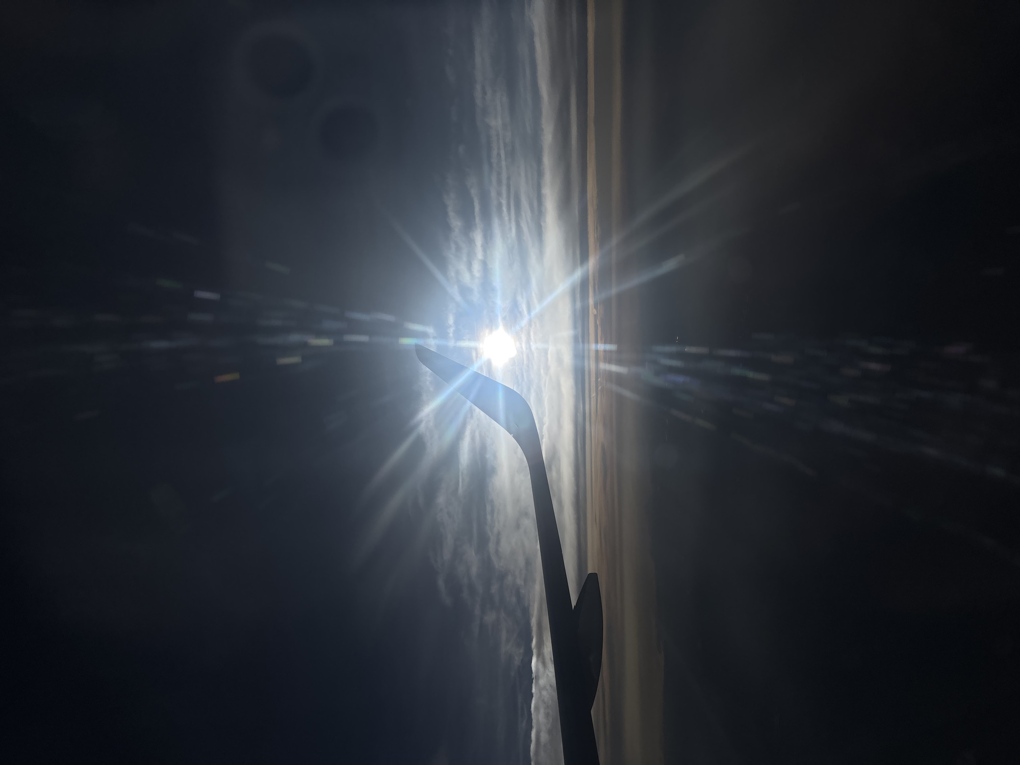 The image shows the sun shining brightly in the sky, partially obscured by the wing of an aircraft. There are clouds and sky visible. The location is inside the cabin of an airplane.