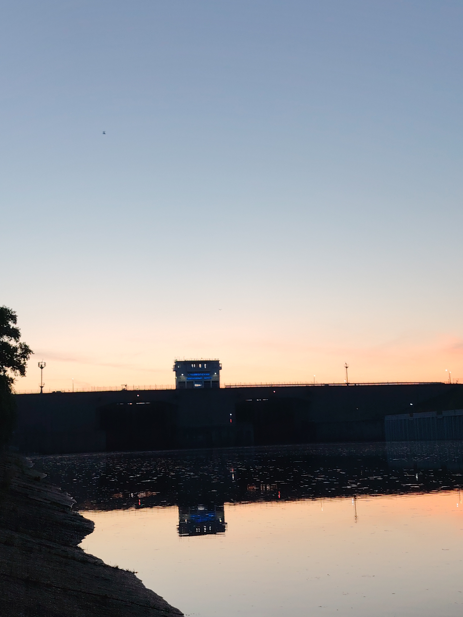 The image depicts a view of the dam in the city of Chaikovsky, Russia at dusk. The sky fades from light blue to orange near the horizon, and the still water of the reservoir reflects the dam structure. The dam is a dark silhouette against the sky, with lit signs in Russian on the control building, and lights lining the top of the structure. A lone tree is visible to the left of the dam. A small bird is flying in the sky. The shoreline is in the foreground on the left, and the water extends to the right, reflecting the colors of the sky and the lights of the dam.