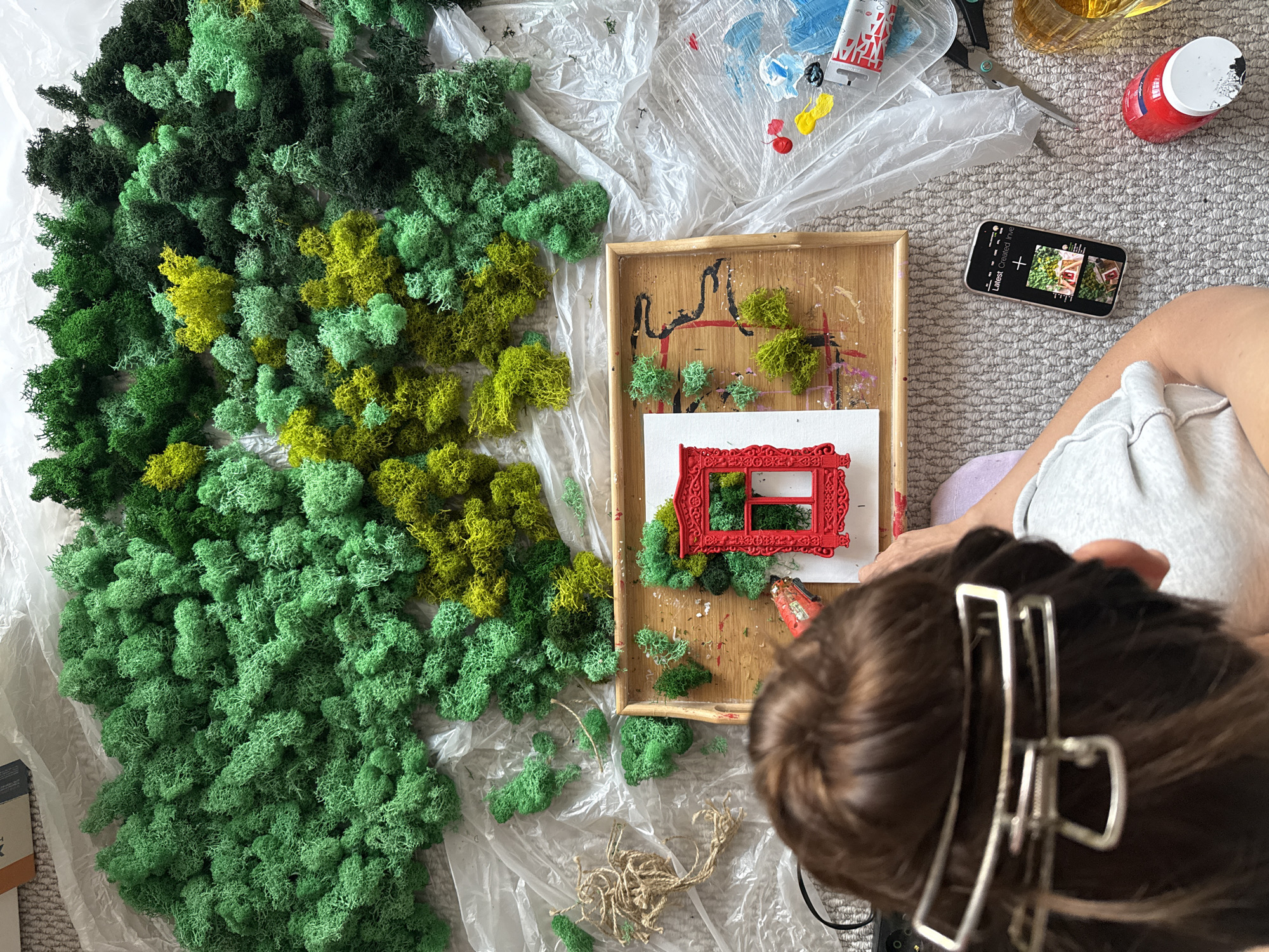 A person is shown from an overhead perspective, seated on a gray carpet. The person has brown hair pulled back with a silver clip. Their left arm is visible, clad in a light gray sleeve, resting on their lap. Before them is a collection of materials, including various shades of green and yellow preserved moss. A wooden tray contains a small red frame, additional pieces of moss, and visible paint splatters. Nearby, there is a smartphone displaying a gallery view of images, a plastic sheet with scattered paint, a tube of paint, a red container, and scissors. The image lacks sufficient information to determine a specific location in Krasnoyarsk, Russia.