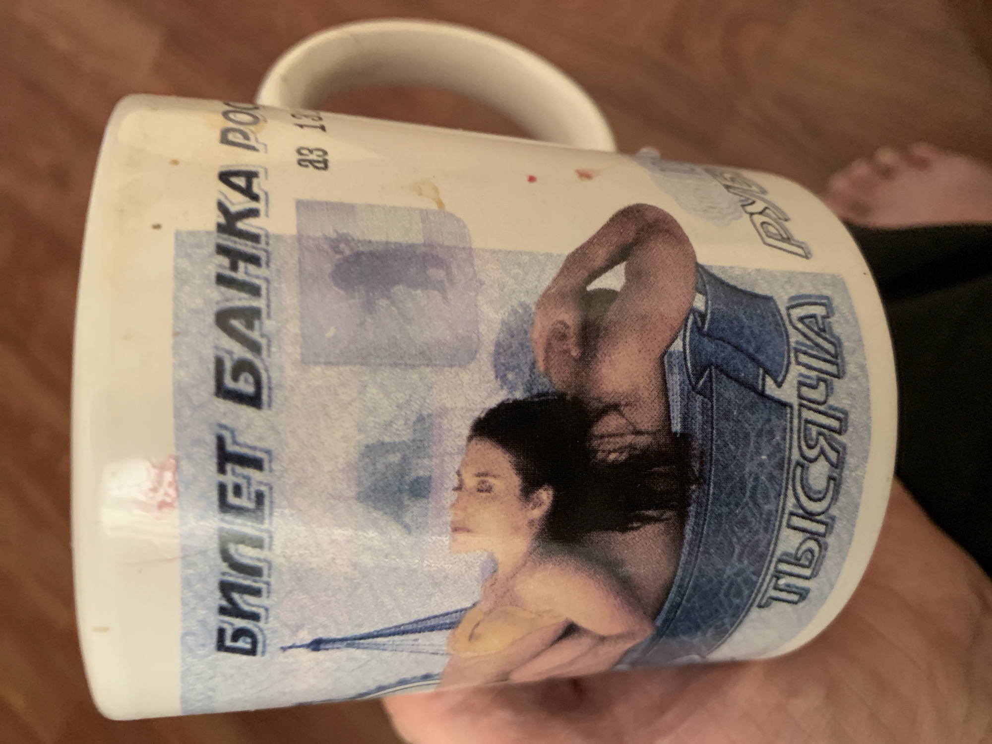 The image shows a white ceramic mug with a design that appears to be based on Russian currency. The design features Cyrillic text, including "БИЛЕТ БАНКА РОСС" (Bank of Russia Ticket) and "ТЫСЯЧА" (Thousand). There is also an image of a woman in a yellow bikini lying down, and a depiction of the Eiffel Tower. The mug has some smudges and marks on its surface. The mug is being held by a person, and their hand and a foot are visible in the frame. The background appears to be a wooden surface, possibly a floor. There is no indication that this image is located in Tashkent, Uzbekistan.