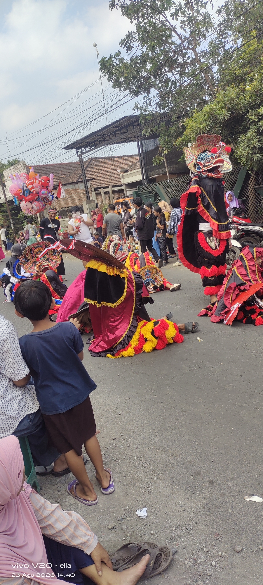In Simo, Indonesia, a street is filled with people watching a traditional performance. The performers are dressed in elaborate costumes, resembling mythical creatures with large, colorful heads and flowing garments. Some performers are on their knees or lying down, while others stand tall. In the background, spectators, including children and adults, are gathered along the street. Balloons and a white truck are also visible. The street is paved, and there are trees and buildings lining the sides. The overall scene depicts a lively cultural event taking place in a public space.