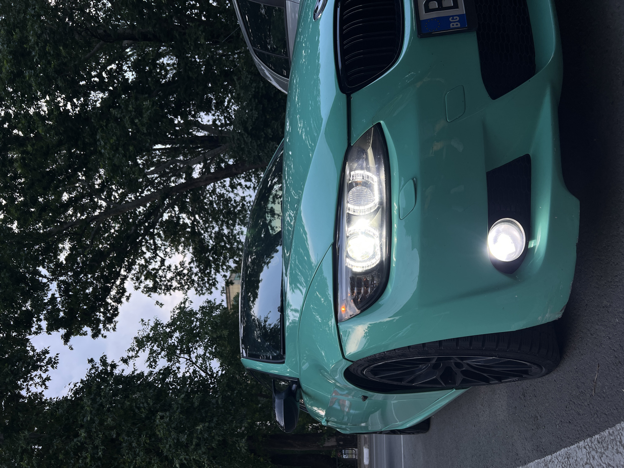 A teal-colored BMW E92/E93 coupe or convertible is parked on asphalt pavement in Asenovgrad, Bulgaria. The vehicle is positioned at an angle, presenting its front fascia and driver's side to the viewer. The car features black multi-spoke wheels, black side mirror caps, and a black kidney grille. Its headlights, including the circular daytime running lights, and the driver's side fog light are illuminated. The driver's side turn signal is also amber-lit. The front bumper shows minor scuffs and imperfections, particularly on the lower left side. A Bulgarian license plate, displaying "BG," is partially visible on the front. A white painted line on the asphalt is visible in the lower left foreground, indicating a parking space or road edge. The background is dominated by a dense canopy of mature deciduous trees, with an overcast or twilight sky visible through gaps in the foliage. A light-colored building and a silver vehicle are partially discernible through the trees on the right side of the frame. No individuals are present in the scene.