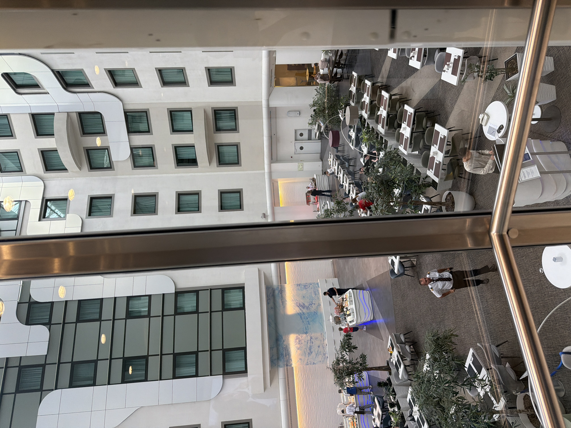The image depicts an interior atrium of a modern hotel, possibly the Novotel Moscow Airport in Khimki, Russia. The atrium is multi-story, with white, curved architectural elements and numerous windows visible on the building's facade. A central courtyard area is furnished with tables and chairs, suggesting a dining or lounge space.
In the foreground, a man in a pilot's uniform, consisting of a white shirt, tie, and dark trousers, walks across the carpeted floor. He is looking down and appears to be heading towards the right side of the frame. A balcony railing with a metallic finish is visible in the lower portion of the image, indicating the vantage point is from an upper floor.
In the middle ground, several people are interacting with a buffet station set up in the atrium. One person, dressed in a black shirt, is attending to the buffet, while a child in a red shirt stands nearby. Another man, wearing a white shirt and apron, is also present near the buffet. Tables are arranged throughout the space, some occupied by diners, others empty. Green potted plants are strategically placed around the atrium, adding a touch of nature to the interior.
The overall ambiance is that of a busy hotel lobby or dining area, with a modern aesthetic characterized by clean lines and a neutral color palette. The lighting appears to be a mix of natural light from the atrium's overhead elements and artificial lighting within the space.