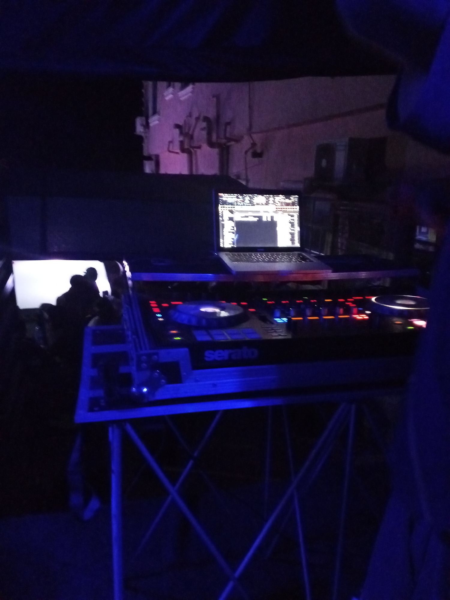 The scene is dimly lit in what appears to be a nightclub or outdoor entertainment venue in Lokoja, Nigeria. Dominating the foreground is a DJ setup consisting of a "Serato" branded DJ controller, a laptop computer, and a stand. Behind the DJ equipment, there is a small screen reflecting light, potentially illuminating the area. Several people are silhouetted in the background, suggesting an audience or crowd. The exterior of a building with pipes and vents is visible behind the DJ setup. The lighting in the scene is primarily blue, casting a cool tone over the entire image.