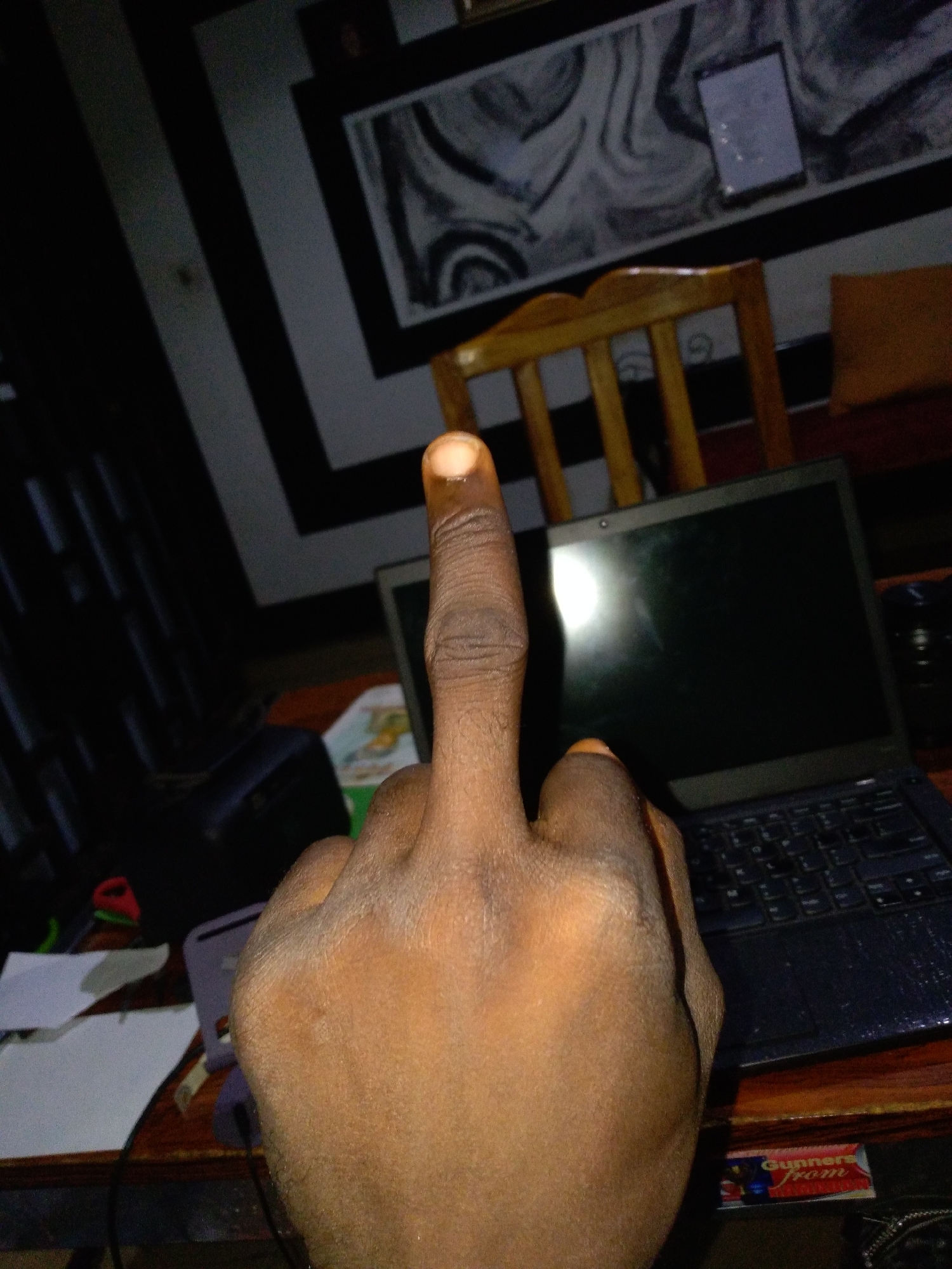 An indoor, close-up shot shows a person's left hand, with the middle finger extended upwards. The person's skin tone is medium brown, and their fingernail is visible and clean. The background is dimly lit, suggesting it is nighttime or the room is dark. A laptop with a black screen is positioned on a wooden desk in the middle ground. To the left of the laptop, a dark blue object, possibly a bag or case, is visible. Further left, on the desk, are papers and what appears to be a red and green object, perhaps a pen or highlighter. In the background, a wooden chair is partially visible, and above it, a wall features a black and white striped pattern and a framed abstract artwork. A small, rectangular item, possibly a phone or wallet, is placed on the striped wall. A product label with the word "Gunners" is visible at the bottom right of the frame. The scene appears to be a desk area in a room, possibly a home office or living space.