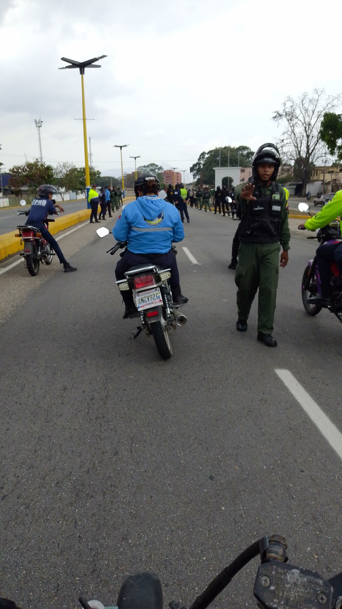 The image shows a scene on a road in Tinaquillo, Venezuela. A person on a motorcycle, with the license plate ANZV92G, is positioned in the center of the road. In front of the motorcycle, a uniformed officer is standing and signaling with their hand. Other individuals, some in uniform and some in civilian clothing, are visible on both sides of the road. Some of the uniformed personnel are on motorcycles. A row of people, likely authorities, are standing along the side of the road near what seems to be a gate or entrance to a structure. There are streetlights on yellow poles along the road. The weather appears to be overcast.