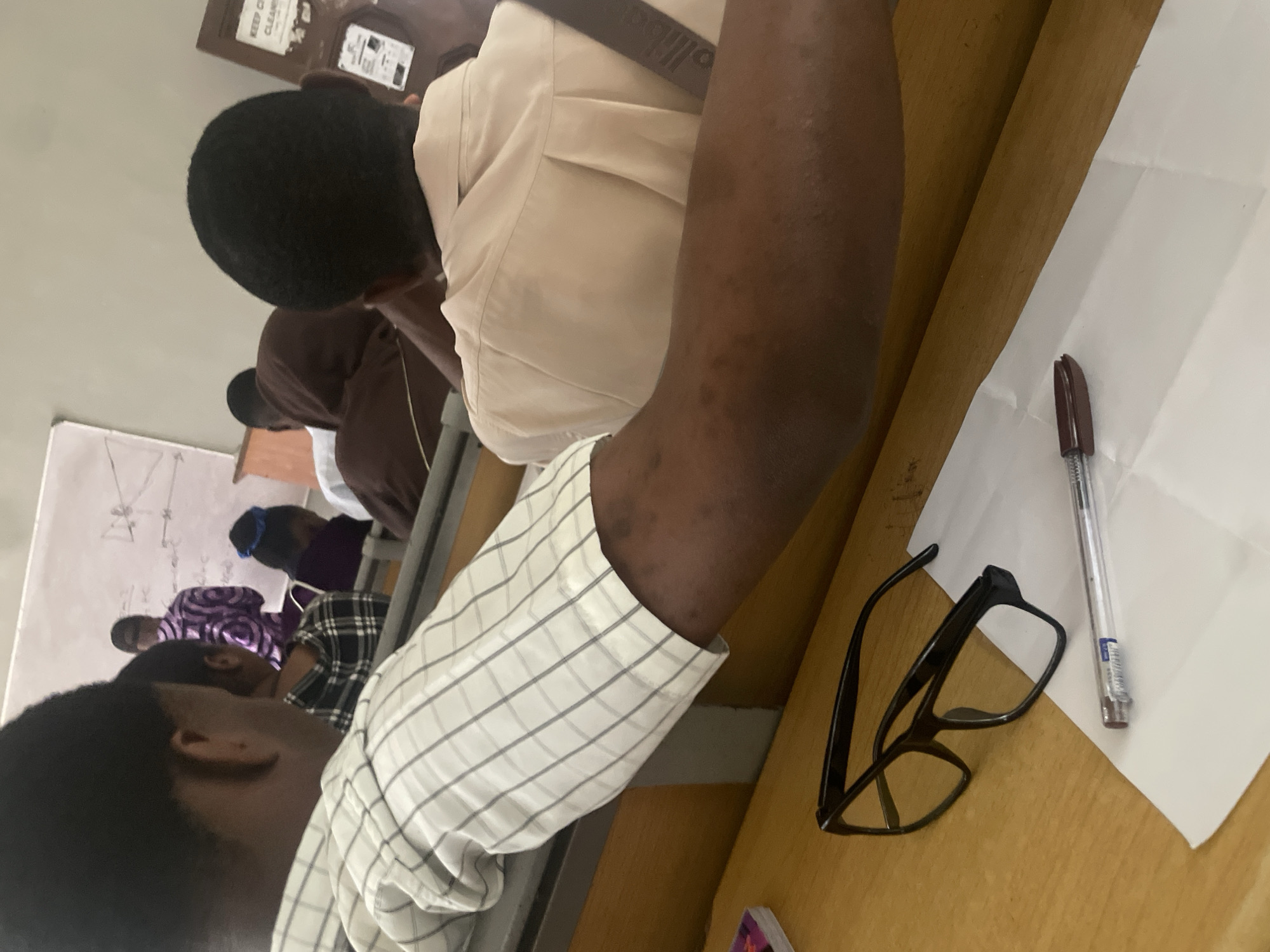 In a classroom setting in Ihiagwa, Nigeria, a student is seated at a desk, their arm resting on the edge of the desk, obscuring much of their upper body. The student is wearing a white and gray checkered shirt. In front of them, also on the desk, are a pair of black-framed glasses and a clear pen with a brown tip, resting on a sheet of white paper. Other students are visible in the background, some seated and facing forward, and one person standing at a whiteboard. The classroom appears to be equipped with wooden desks and chairs, and a projector screen or whiteboard is mounted on the wall. The lighting suggests an indoor environment.