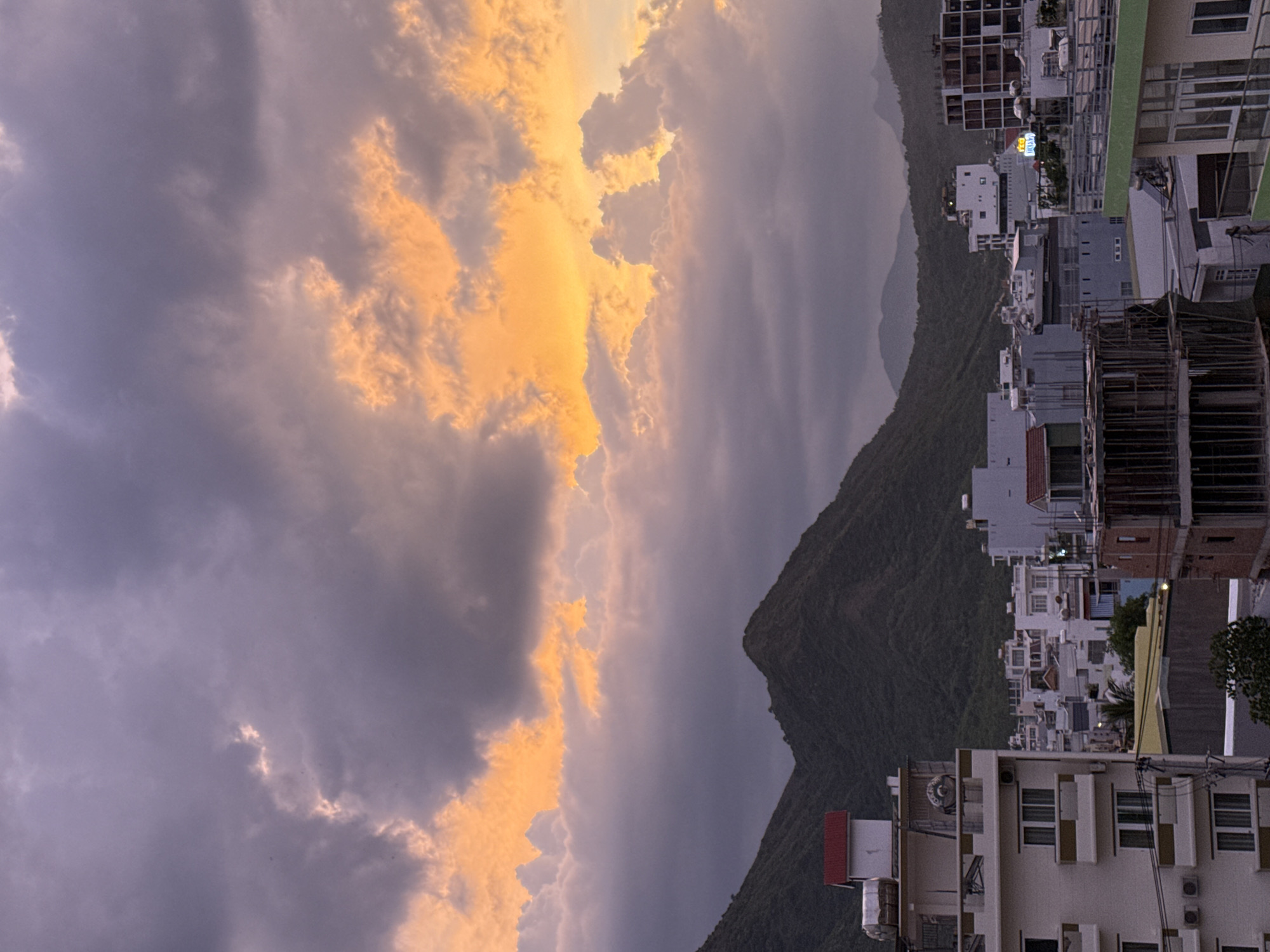 The image depicts a coastal city in Nha Trang, Vietnam, during what appears to be sunset or sunrise. The sky is filled with dramatic clouds, displaying hues of purple, grey, and vibrant orange, suggesting the sun is low on the horizon. In the foreground, several multi-story buildings are visible, some appearing to be under construction, with exposed rebar and concrete frameworks. Other buildings are completed, featuring balconies, windows, and air conditioning units. A prominent red-roofed structure sits atop one of the buildings. Behind the city, lush green mountains rise, their slopes covered in vegetation. The overall scene conveys a sense of an urban environment nestled against a natural landscape, bathed in the warm, ethereal light of twilight. There are no people or animals visible in the frame.