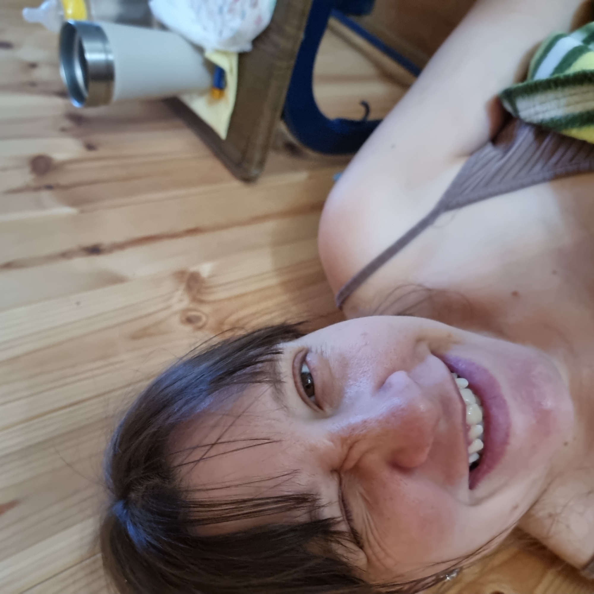 The image shows a close-up, selfie-style shot of a person's face and upper body. The person is winking and smiling, with their mouth open to reveal teeth. They have dark hair and are wearing a thin-strapped top. Behind them, the background consists of light-colored wooden paneling. On a surface to the right, there is a white travel mug with a silver rim, a yellow object, and what appears to be a baby bottle and some white fabric. The lighting is warm and suggests an indoor setting. There is no indication of the specific location within Dunnae-myeon, South Korea.