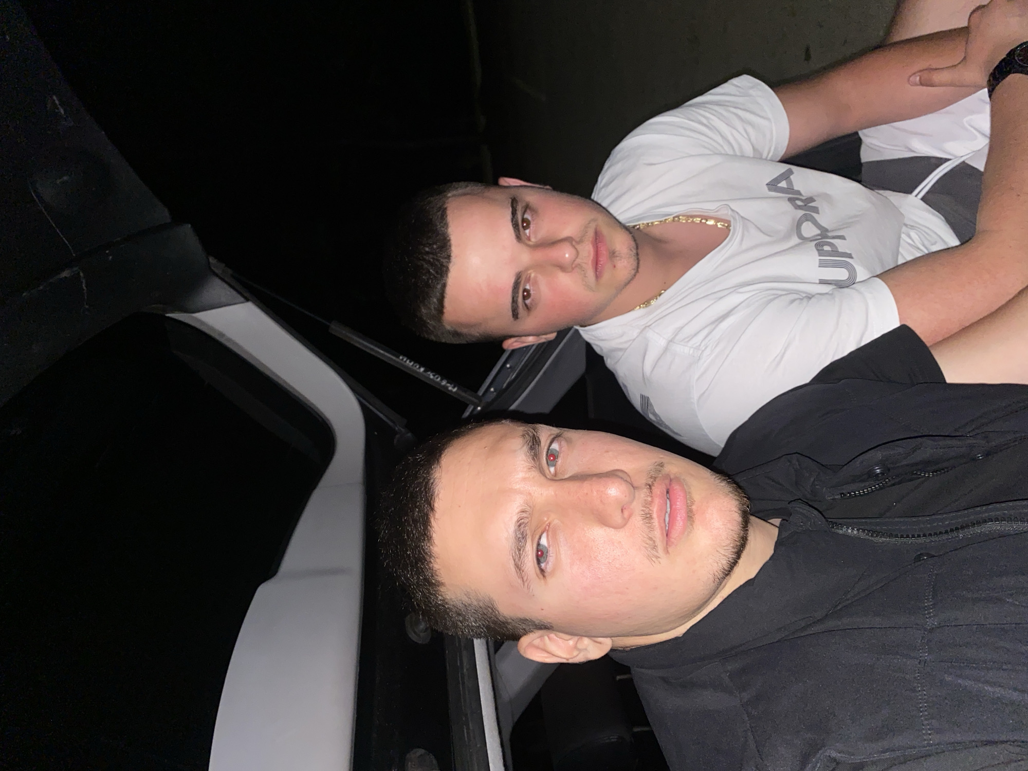 In the city of Pleven, Bulgaria, two young men are seated in the open trunk of a vehicle at night. The man on the left, wearing a black vest over a black t-shirt, has short dark hair, a thin mustache, and a goatee. His eyes appear red, possibly due to the flash of the camera. The man on the right, wearing a white t-shirt with a grey graphic and a gold chain, also has short dark hair and a light beard. Both men are looking directly at the camera with neutral expressions. The background is dark, suggesting an outdoor setting at night. The vehicle's interior, including seats and the trunk lid, is visible behind them. The overall scene is candid and casual.