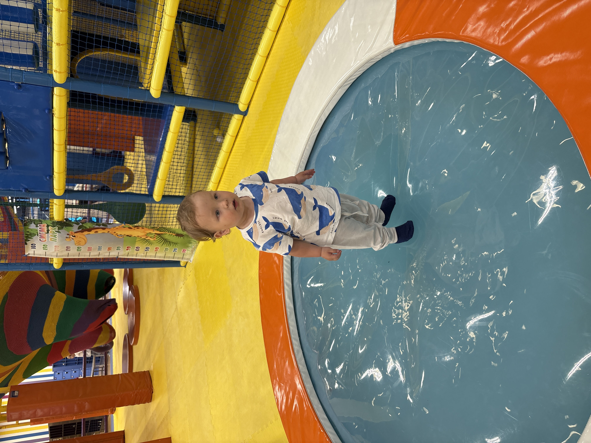 A toddler stands in a circular play area with a blue floor covered by a clear plastic sheet, bordered by white and orange cushioned edges. He is wearing a white t-shirt with blue whale designs, light gray pants, and dark blue socks. The surrounding environment is a colorful indoor playground with yellow flooring, blue and yellow netting, and other play structures. A giraffe-themed height chart is visible on a wall. The photo appears to have been taken at a children's entertainment center in Izhevsk, Russia, named "Siti Park."