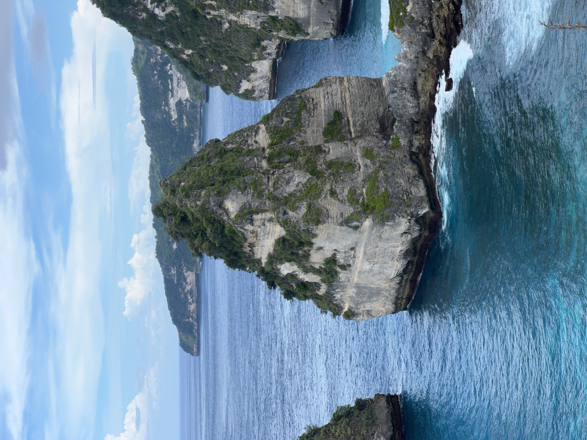 This media file captures a dramatic coastal landscape in Nusa Penida, Indonesia, presented in a vertical orientation under bright daytime conditions with a clear sky. The left side of the frame is dominated by a bright blue sky with scattered white clouds.
The central and right portions of the image showcase majestic, rugged limestone cliffs and towering sea stacks rising sharply from the vibrant ocean. A prominent sea stack, covered with lush green tropical vegetation, stands centrally, its steep, grey-white rock walls plunging into the water. Towards the upper part of the image, to the left of this central formation, a deep blue inlet or bay stretches back, bordered by verdant cliffs, with more distant landmasses fading into the background.
Towards the lower part of the image, the ocean transitions from deep indigo to a bright turquoise, especially where white, foamy waves are visibly breaking and crashing against the bases of the rock formations. Part of another vegetated rocky outcrop is visible in the bottom right corner, also experiencing wave action.
The varied shades of blue in the water, the ancient grey-white rock, and the vibrant green foliage create a striking contrast. No human subjects, man-made objects, or visible text are present, allowing the untouched natural grandeur of the landscape to be the sole focus. The image conveys a profound sense of awe and unspoiled beauty.