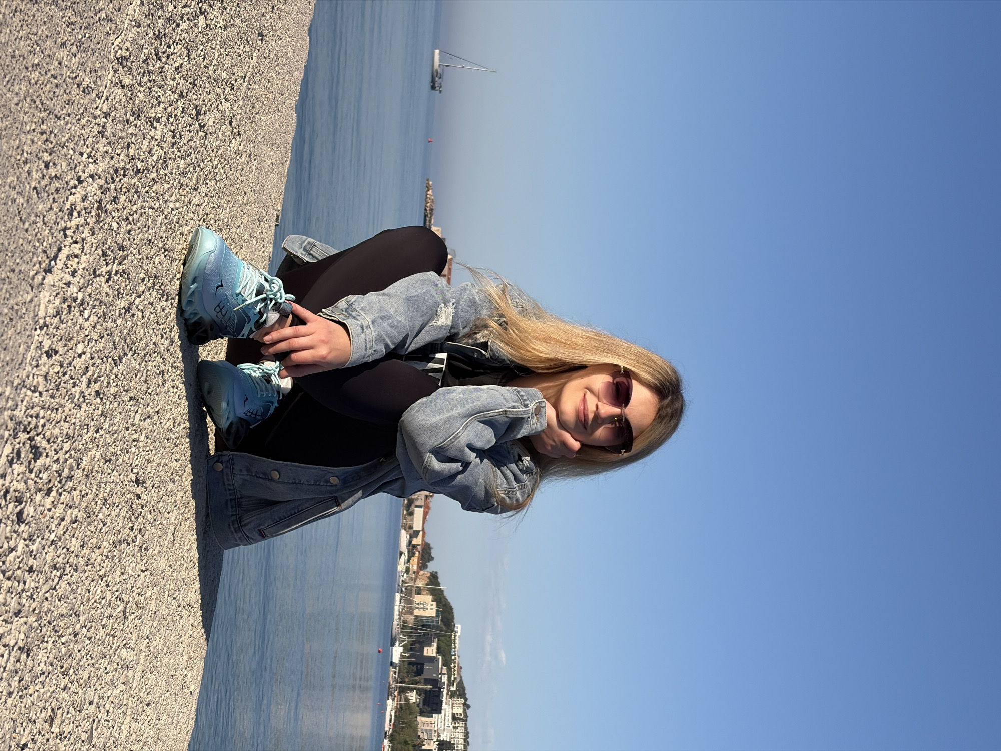 A blonde-haired woman wearing sunglasses, a denim jacket, and black leggings is sitting on a stone structure next to a body of water. She is wearing blue sneakers. In the background, there is a cityscape and a sailboat on the water. The location appears to be in Budva, Montenegro, given the coastal urban setting and architectural style. The sky is blue and clear. The woman is resting her face on her hand and looking at the camera.