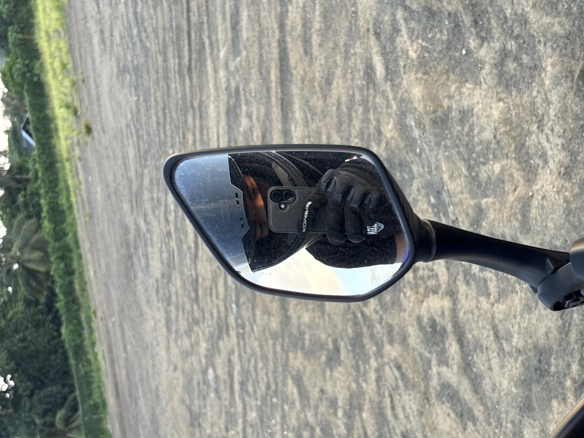 The image is a first-person perspective, likely taken by someone riding a motorcycle, showing a reflection in a motorcycle mirror. The reflection captures a person wearing a black helmet and black gloves, holding a smartphone to take the picture. The background of the reflection shows a glimpse of a person's black t-shirt with white text. The environment outside the mirror is a sandy or dirt road with sparse vegetation in the background, possibly leading to a beach or coastal area. The presence of palm trees and dense green foliage suggests a tropical location. Based on the vegetation and general landscape, this scene could be situated in a coastal or semi-rural area of Davao City, Philippines, known for its tropical climate and natural landscapes. The lighting suggests it is daytime, possibly late afternoon given the angle of the light.